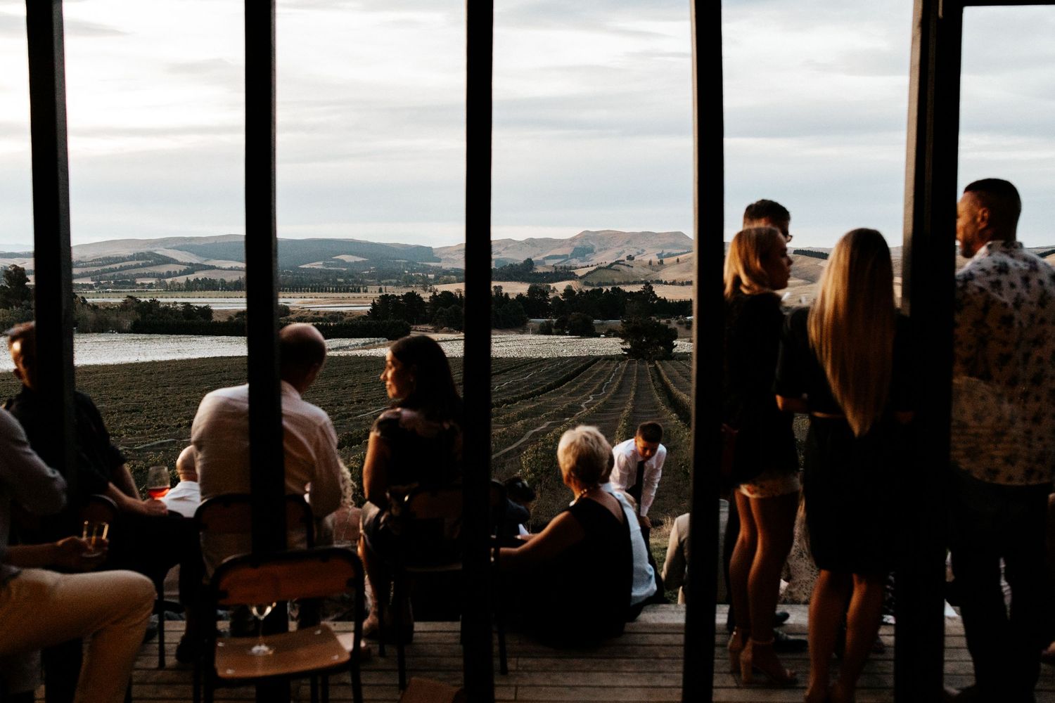 Wedding guests gather by large windows overlooking a scenic mountain landscape.