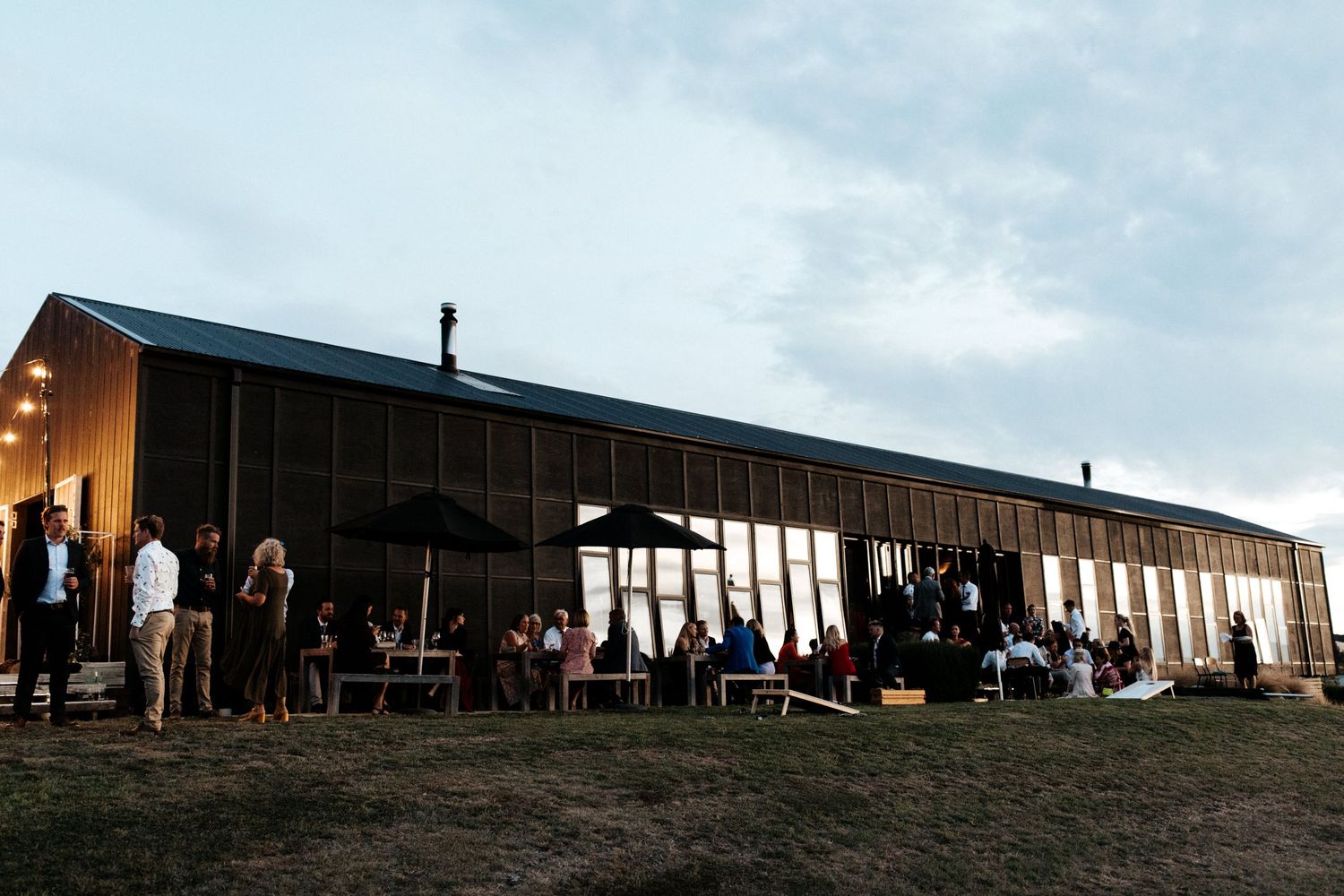 Modern barn wedding venue at dusk with string lights and guests gathered outside.