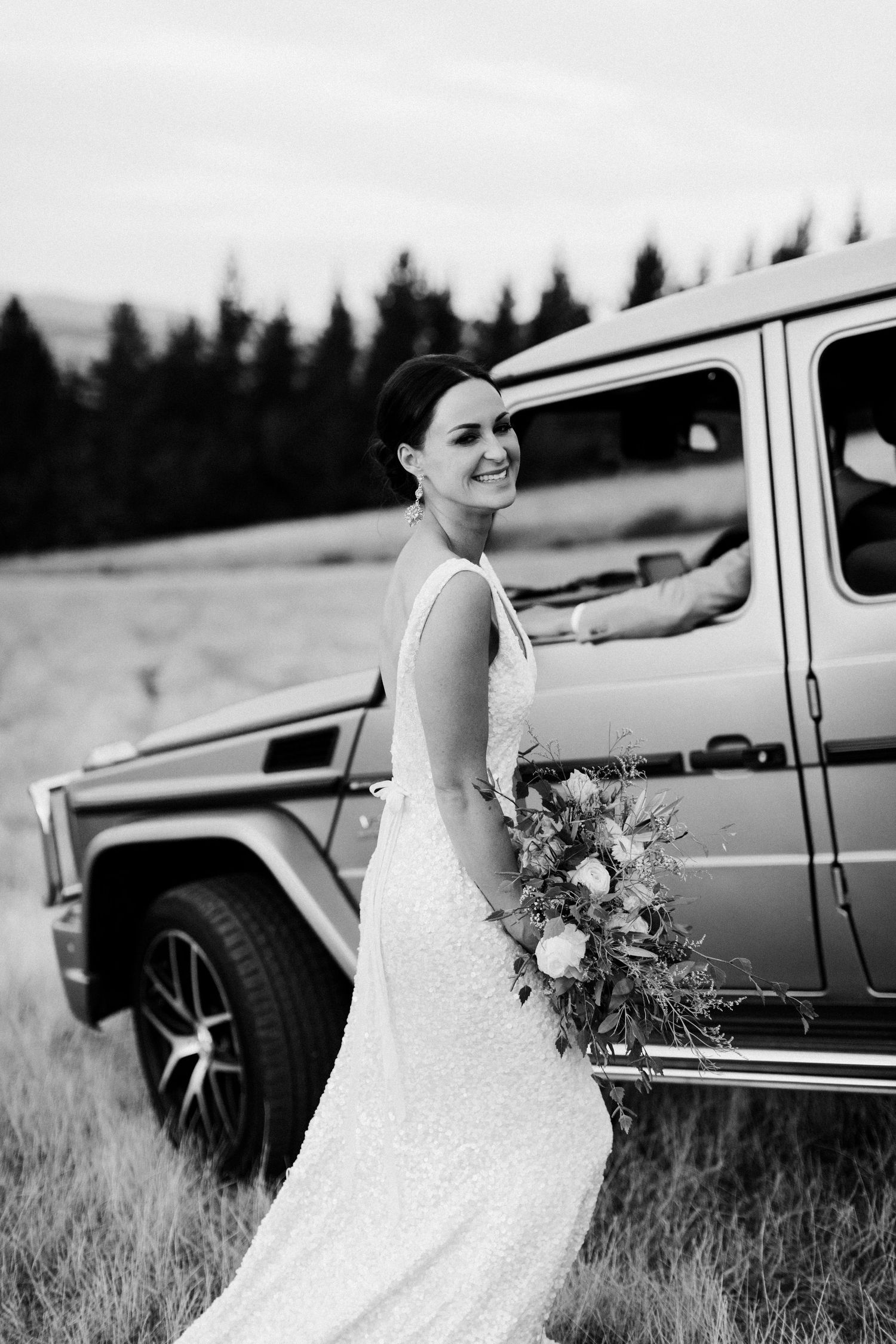 A bride in a fitted white dress poses with a silver G-wagon vehicle in a field with mountains in the background.