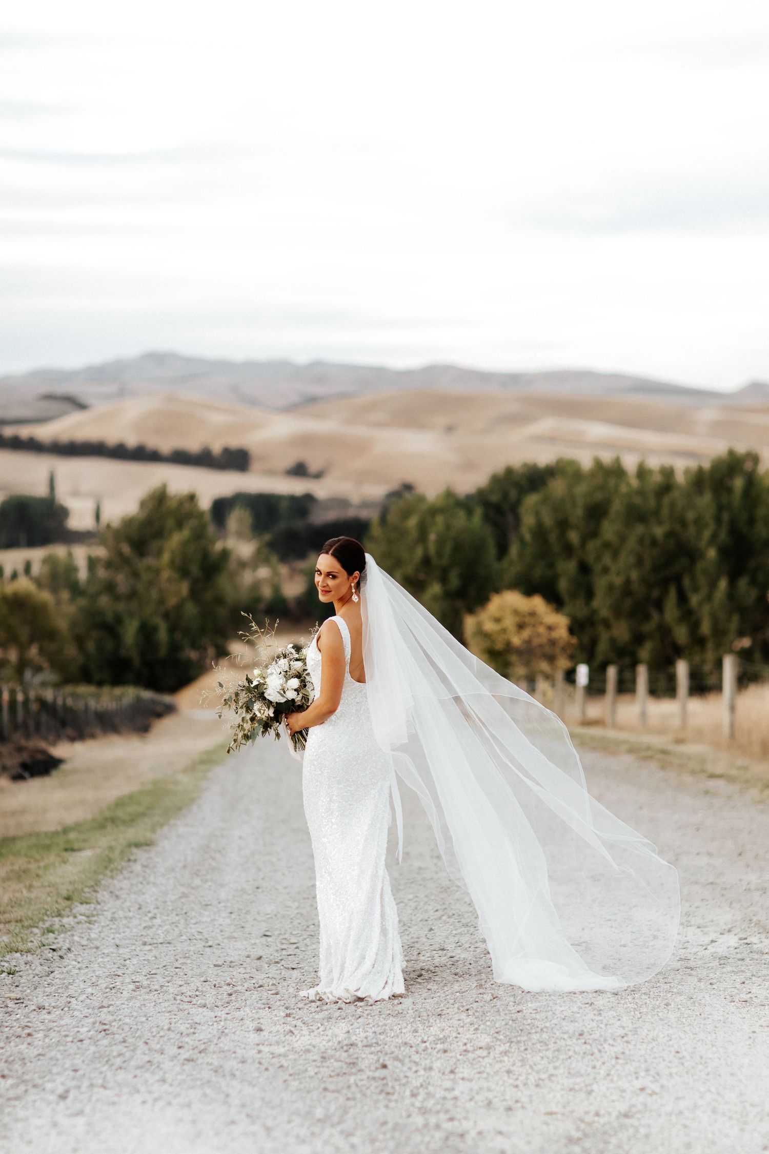 Bride with flowing veil poses on country road with scenic mountain backdrop.