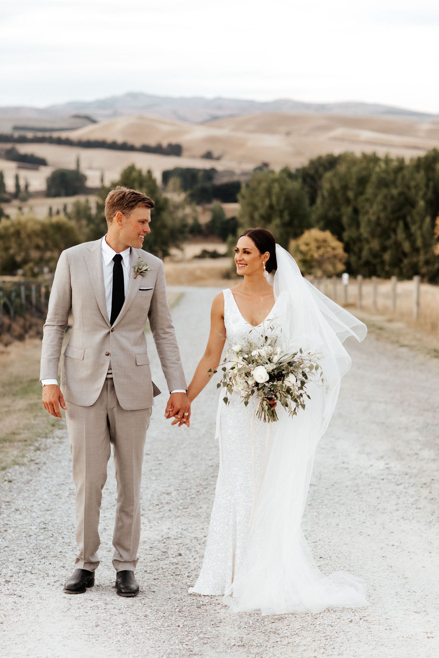 Elegant couple walks hand in hand on gravel path with rolling hills in background.