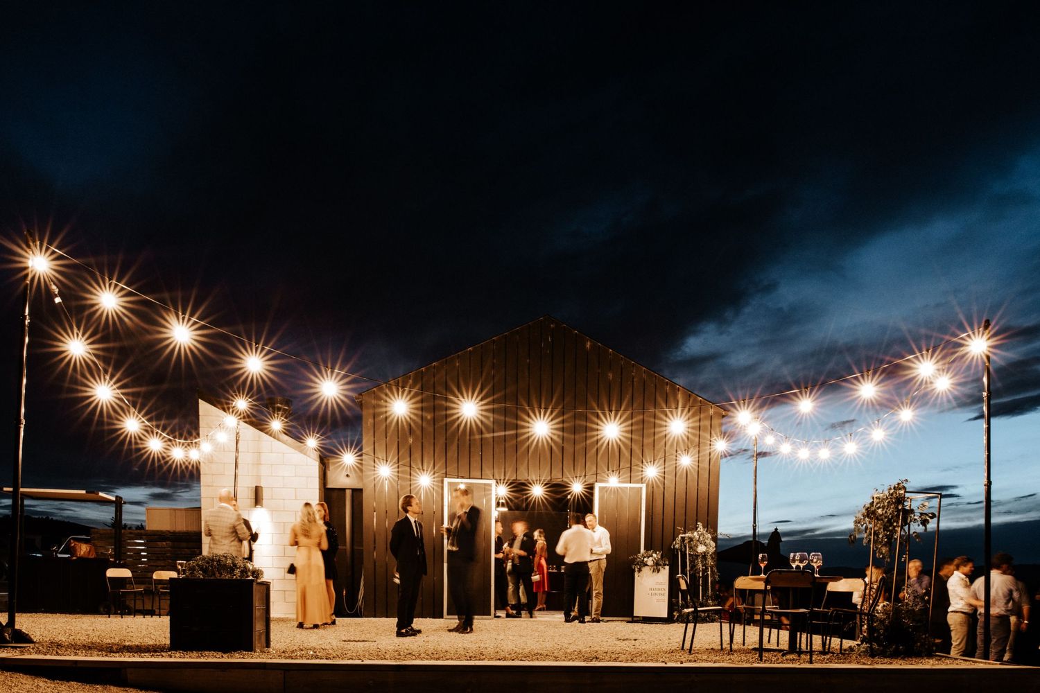 String lights illuminate an outdoor venue at dusk with guests gathered for an evening event.
