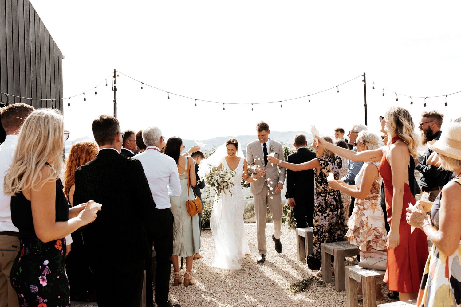 Wedding guests gather under string lights to celebrate the newlyweds at an outdoor reception.