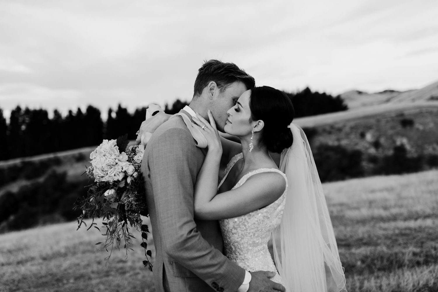 Black and white photo of a romantic wedding kiss with bouquet of flowers against mountain backdrop.