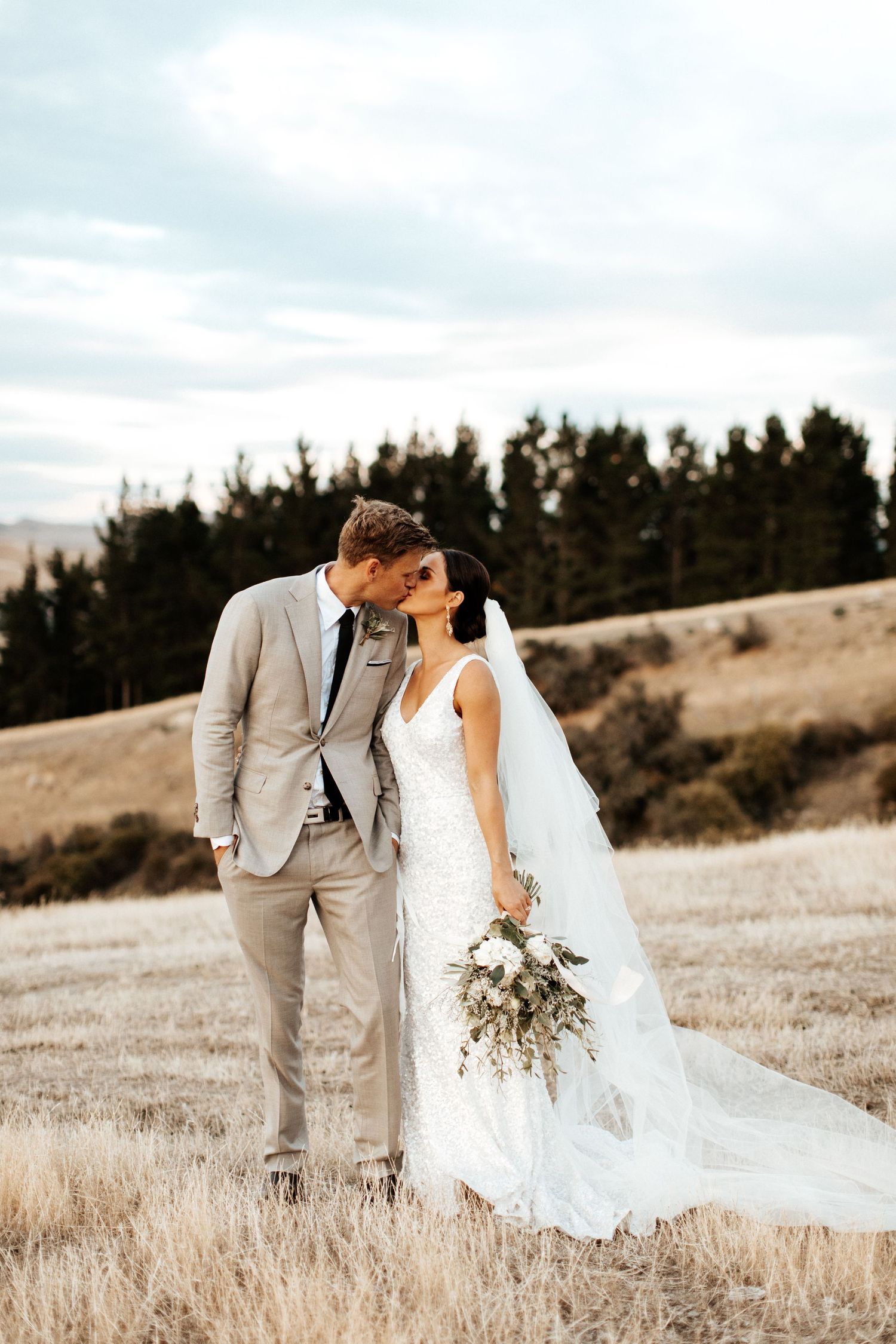 Bride in flowing white gown and groom in light grey suit embrace on scenic hillside.