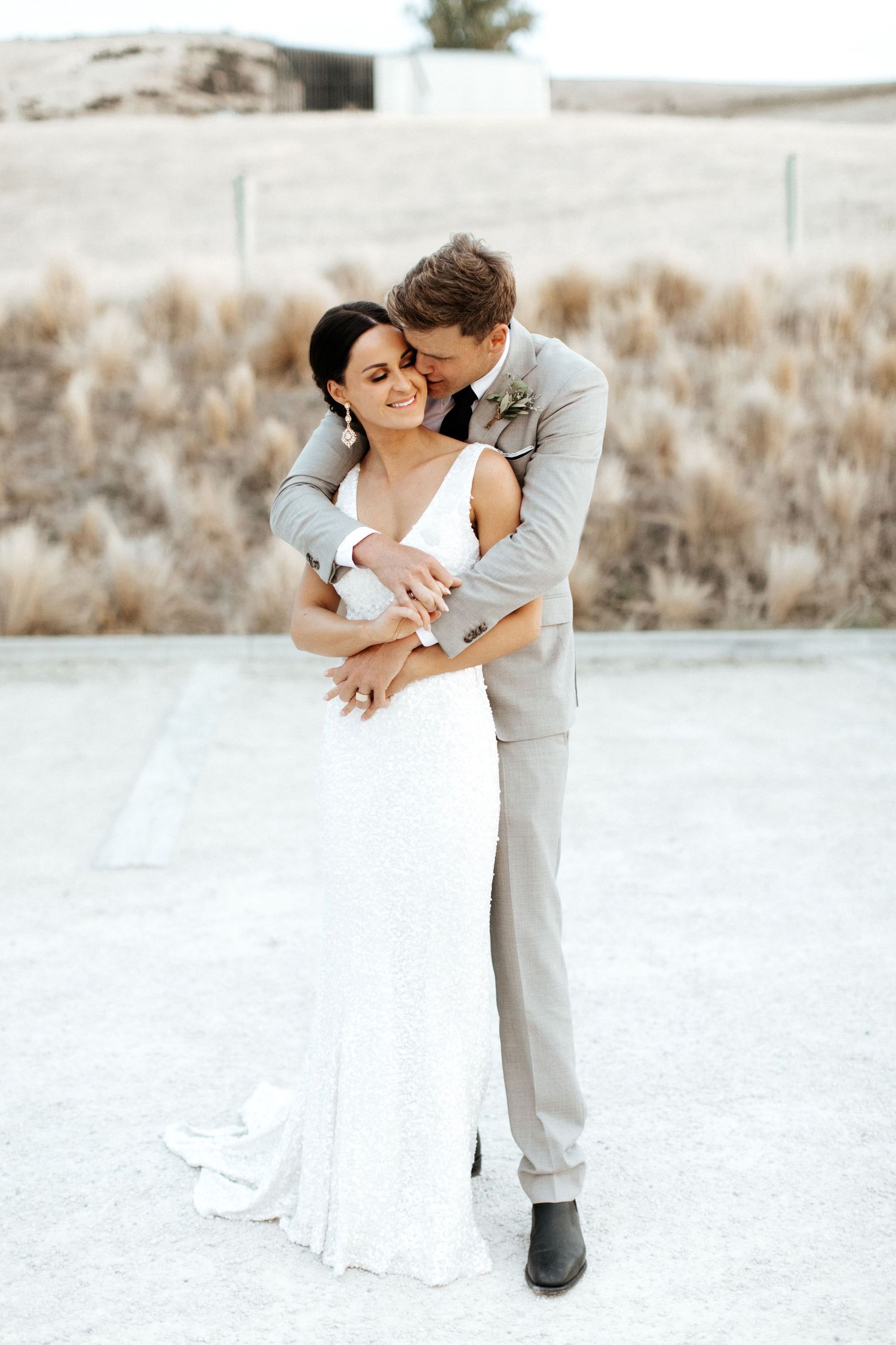 Newlyweds share an intimate moment against desert backdrop in elegant wedding attire.