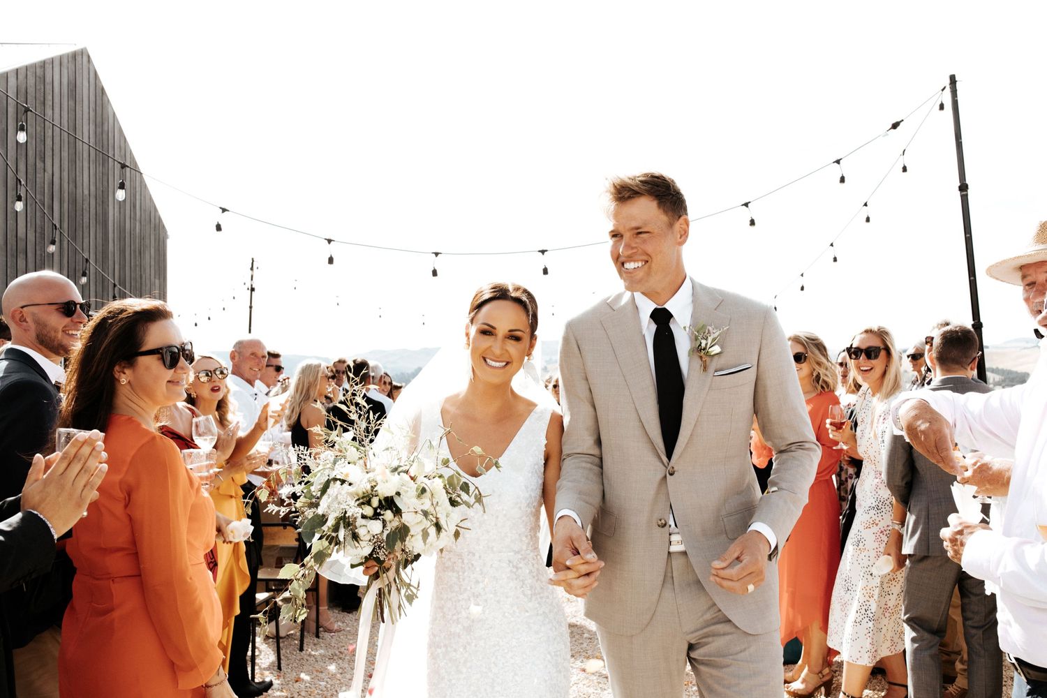 Newlyweds walk through cheering guests under string lights at their outdoor wedding reception.