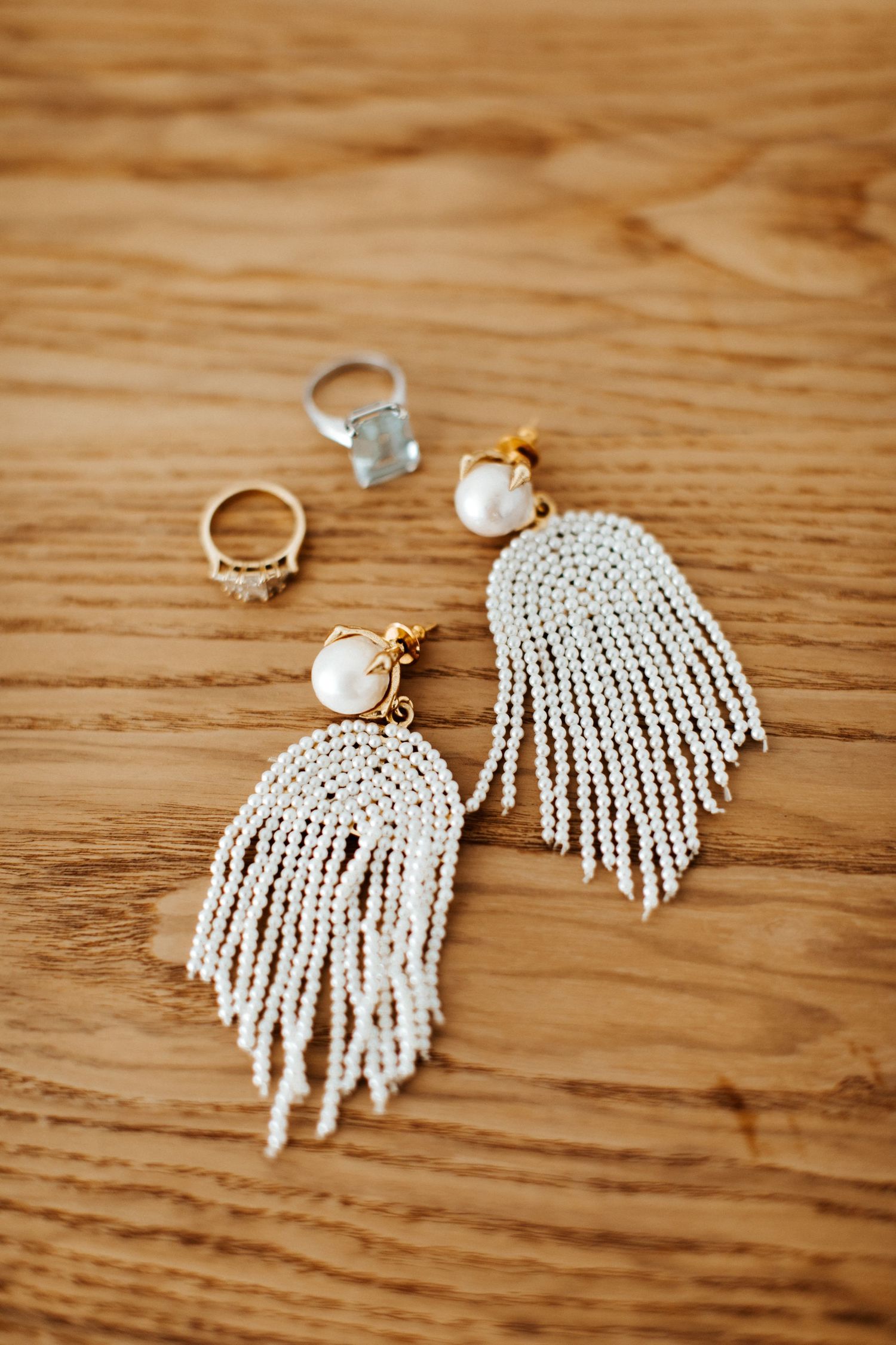 Pearl beaded fringe earrings and rings displayed on wooden surface for wedding accessories.