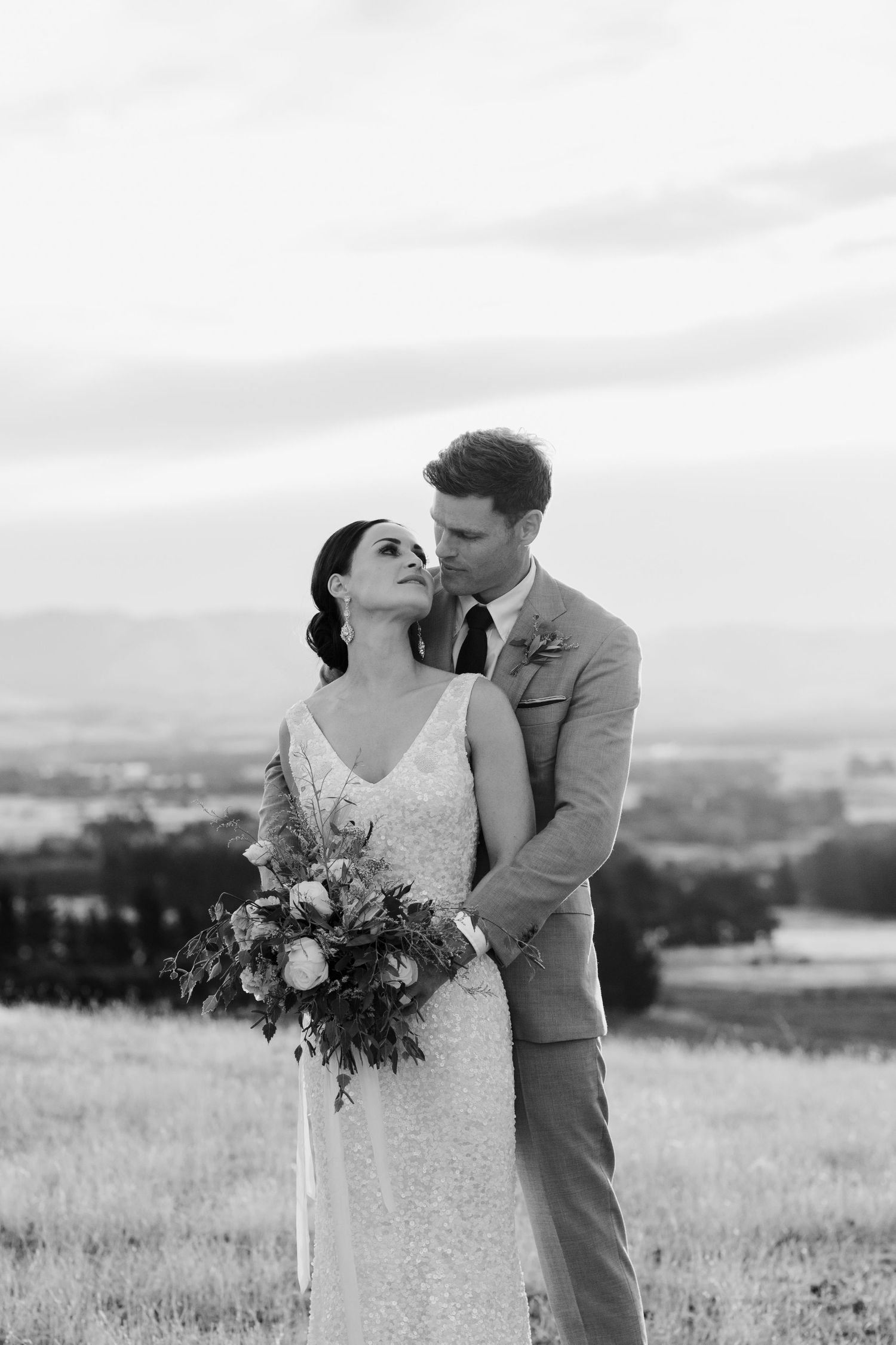 Black and white portrait of wedding couple embracing on scenic hillside with mountain views.