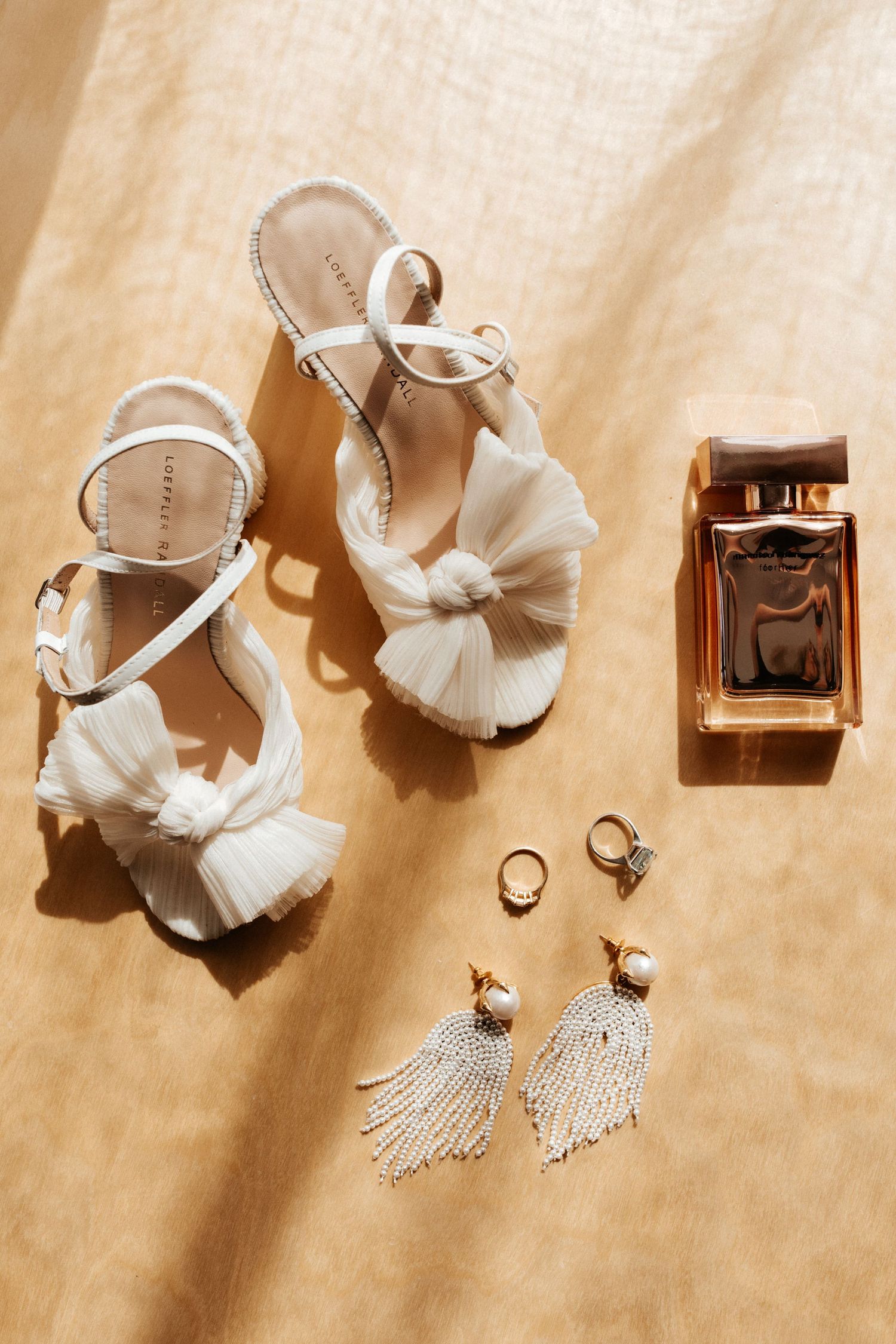 Wedding accessories including white sandals, perfume bottle, and dangling earrings on beige fabric.