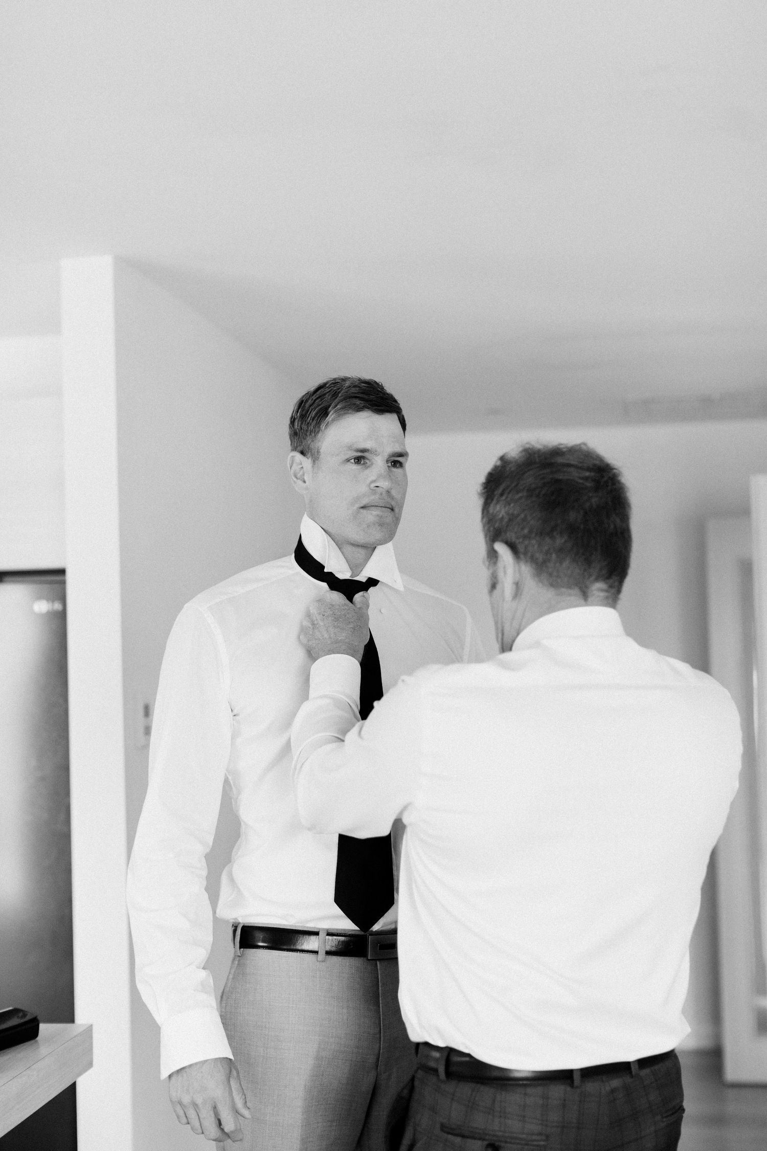 Men in formal attire adjusting a tie during wedding preparations.