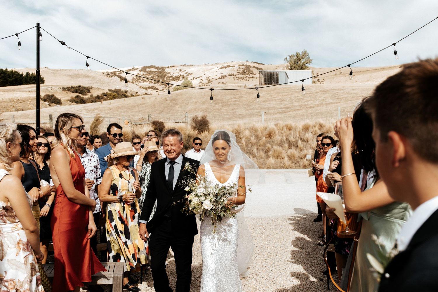 Wedding ceremony with guests watching couple walk down aisle under string lights.