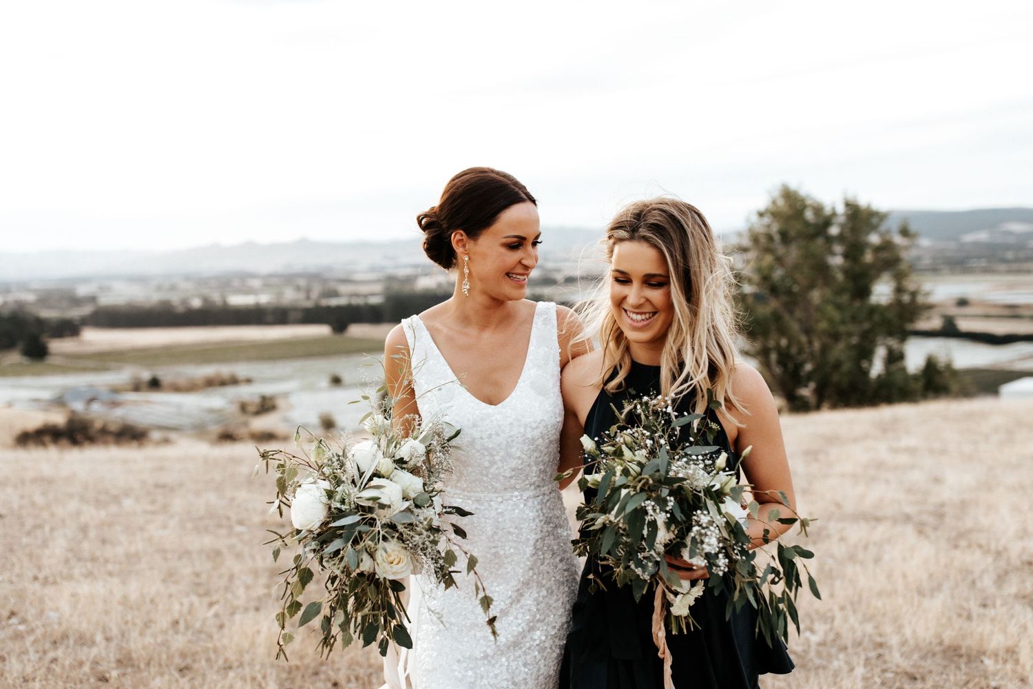 Wedding party members smile while holding matching white and green bouquets outdoors.