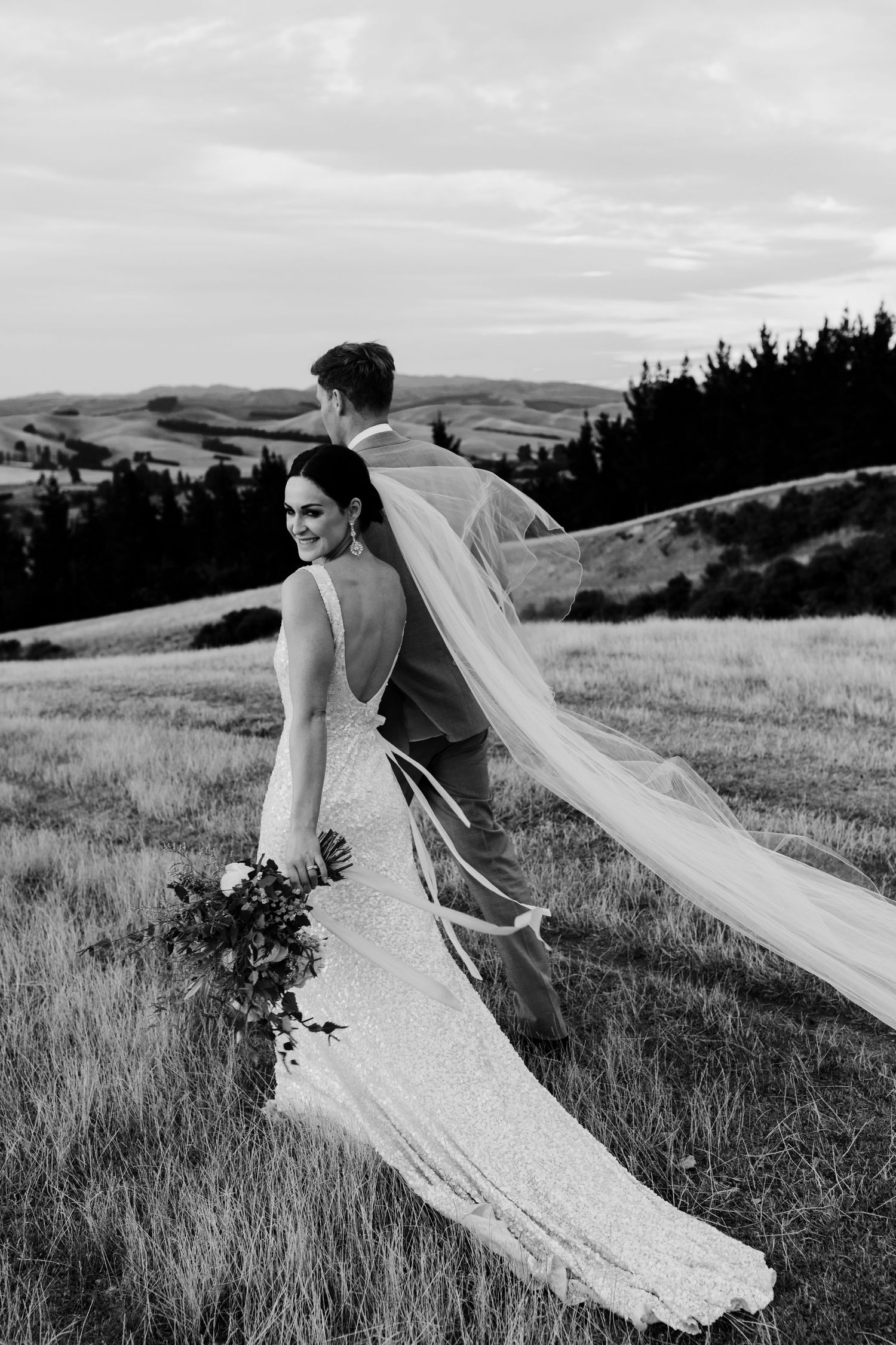 Black and white wedding photo with flowing veil creating dramatic silhouette against pastoral landscape.
