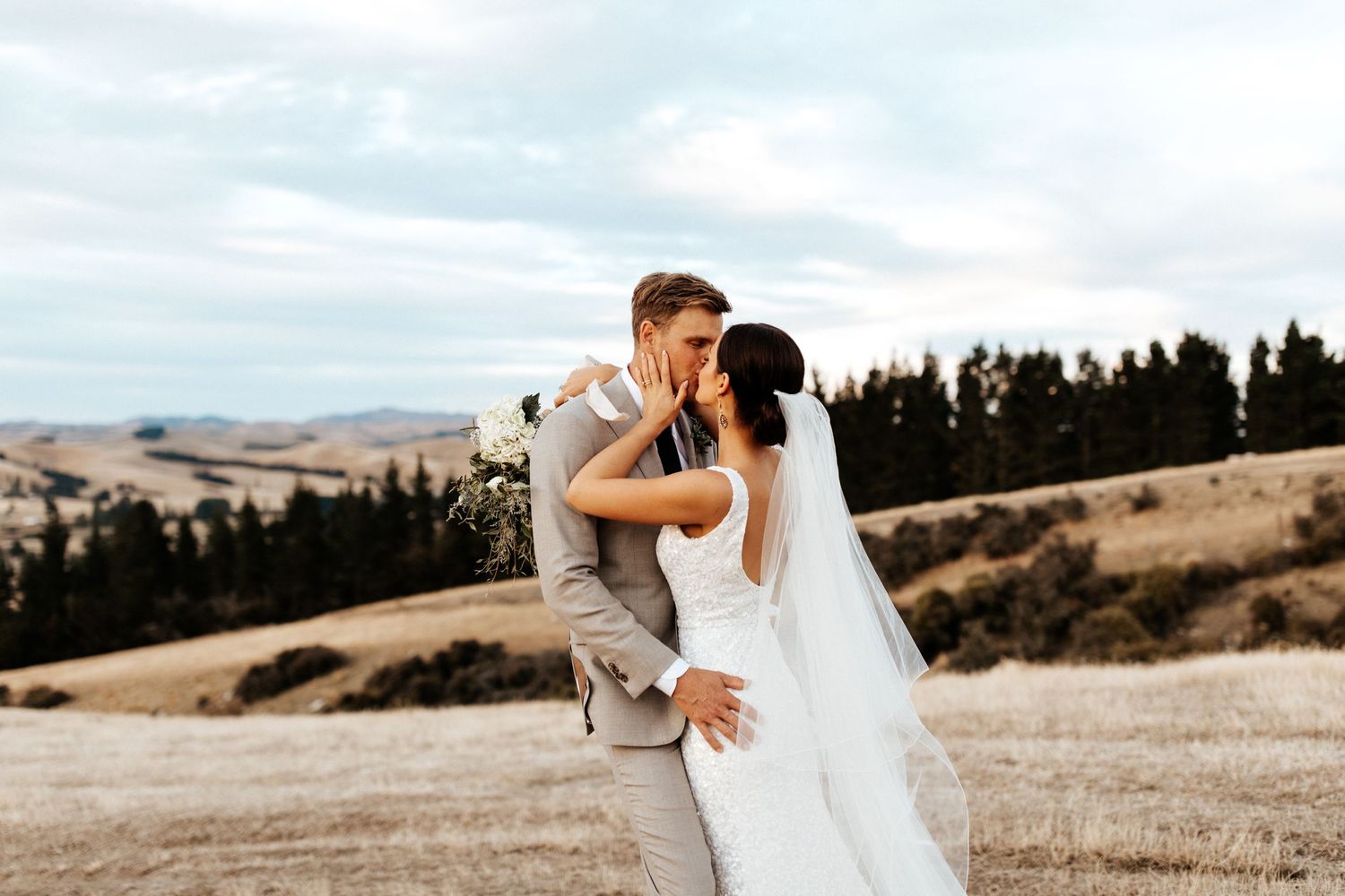 Newlyweds share an intimate kiss on a hilltop overlooking rolling hills and evergreen trees.