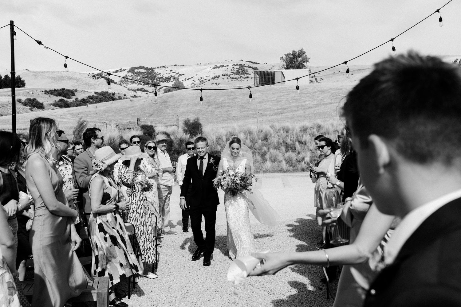 Black and white shot of wedding ceremony processional with guests lined up outdoors.
