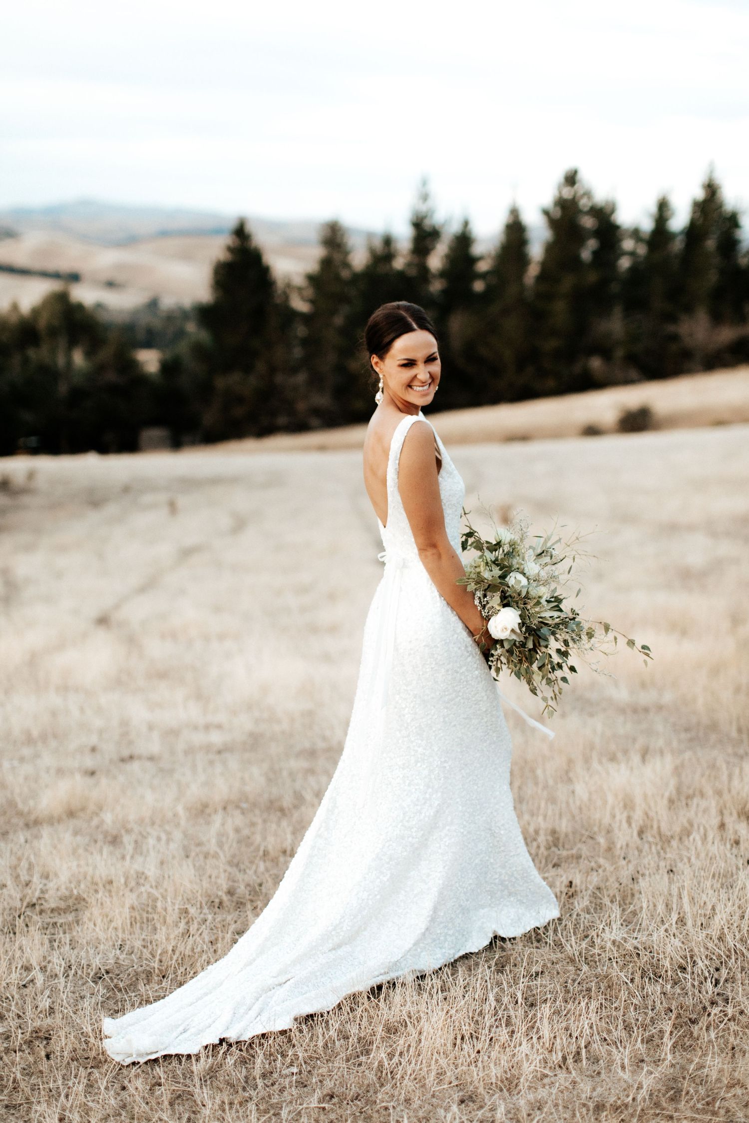 Bride in white gown holds bouquet while standing in sun-dried grass field.