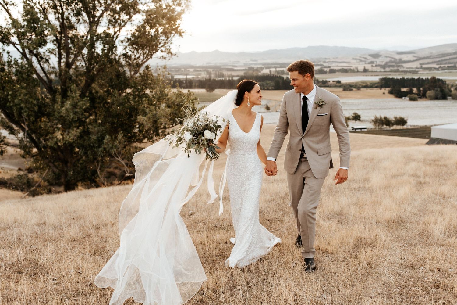 Wedding couple walking through golden grass on hillside with flowing white veil and bouquet.