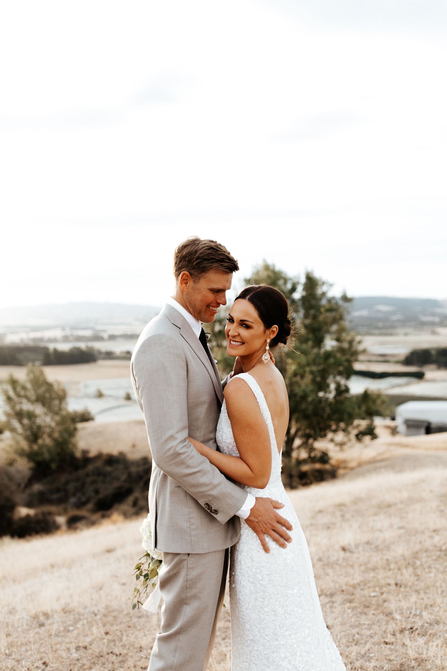 Romantic portrait of couple in wedding attire embracing on hill with countryside backdrop.