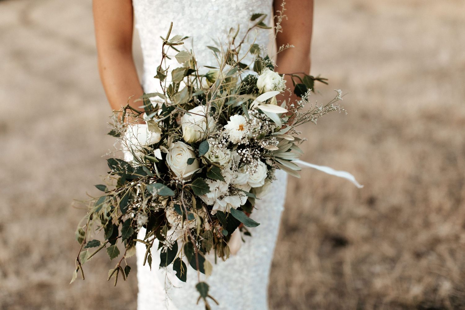 Rustic white and green wedding bouquet with flowing ribbons held against lace dress.
