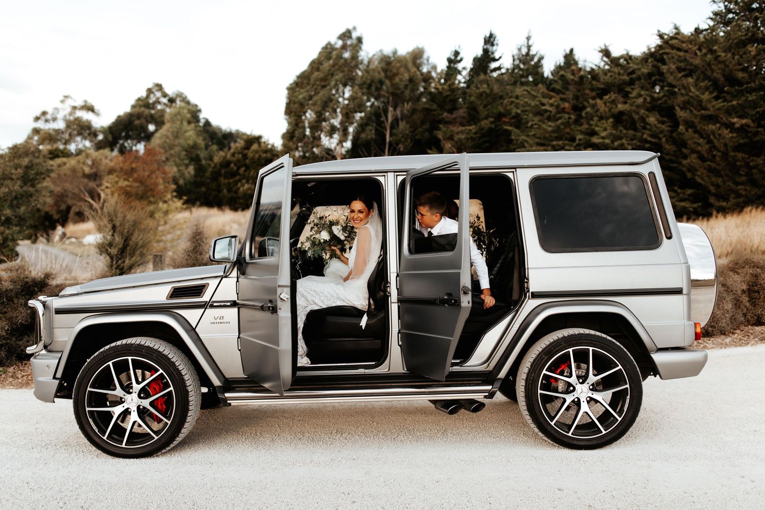 Wedding party poses in a luxurious silver Mercedes G-Wagon with doors open.