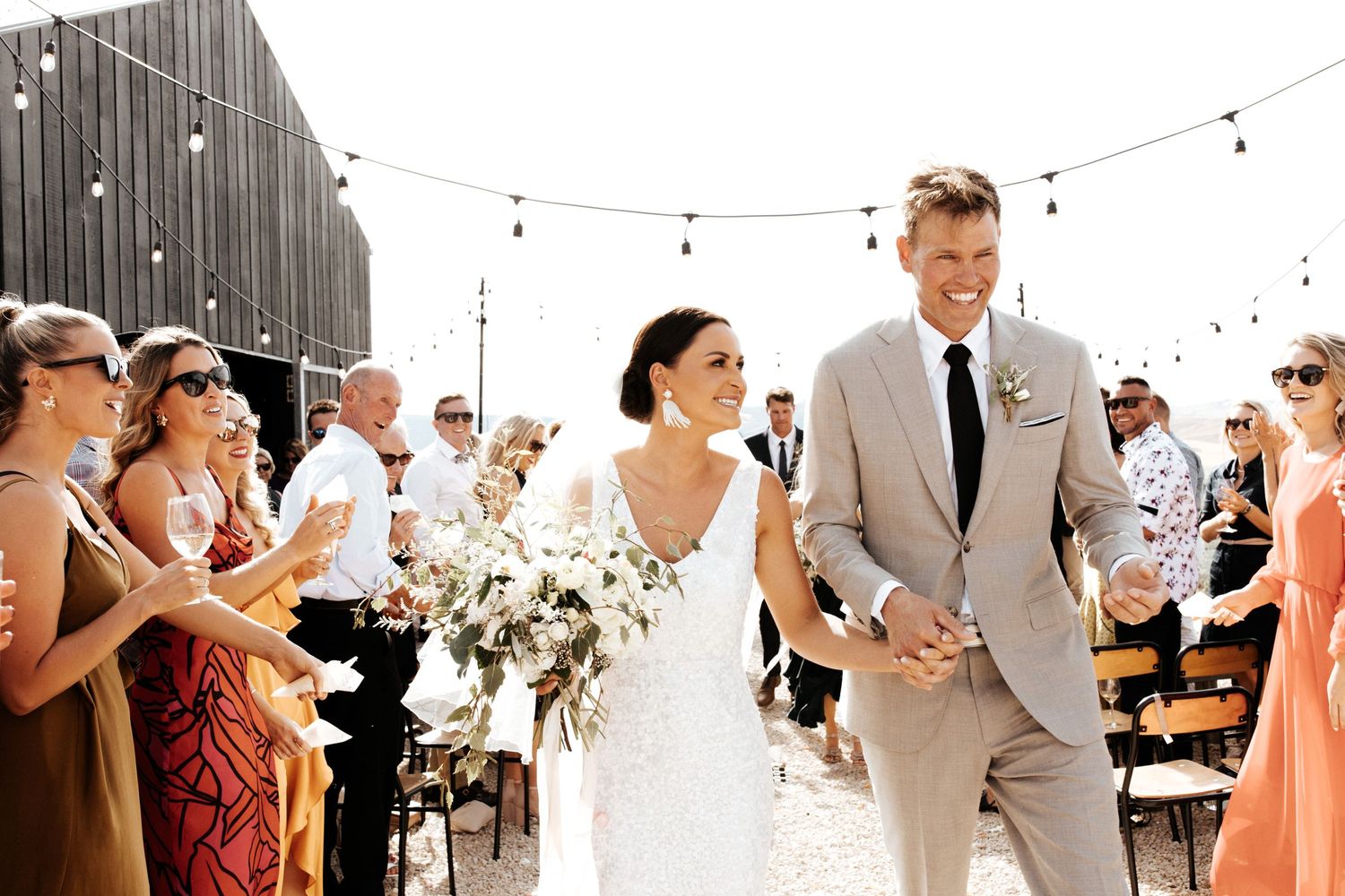 Happy couple makes their grand exit as wedding guests cheer at an outdoor venue with string lights.