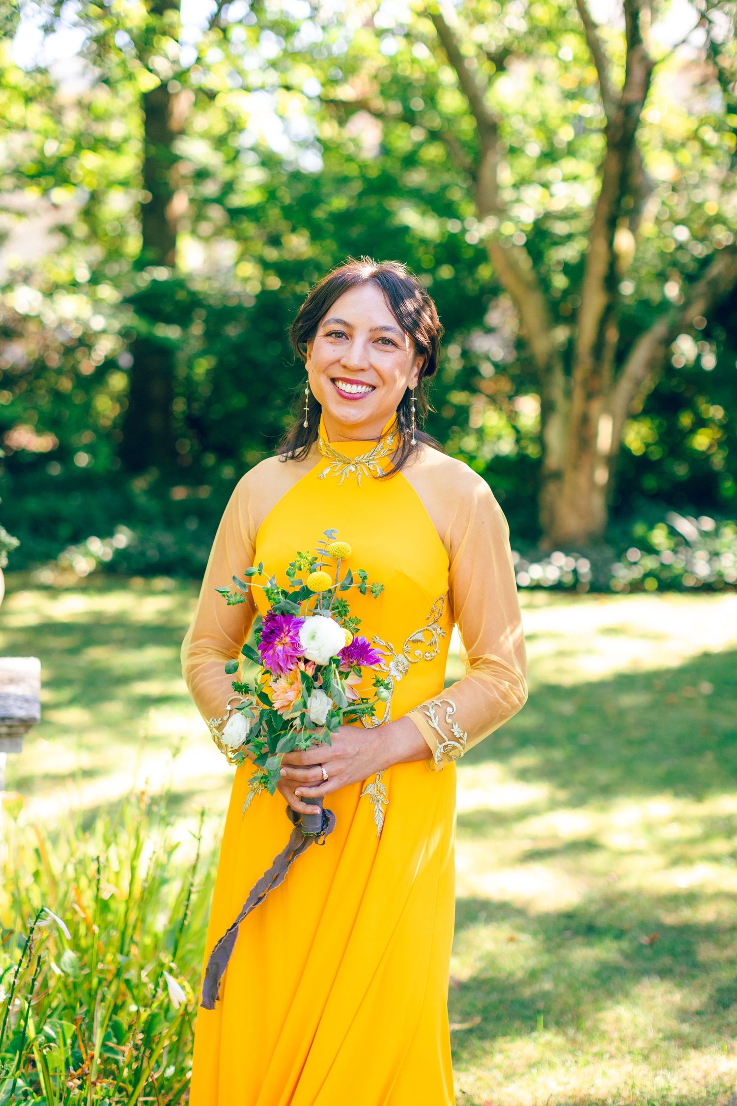 Woman in yellow dress holds colorful bouquet while standing in sunlit garden.