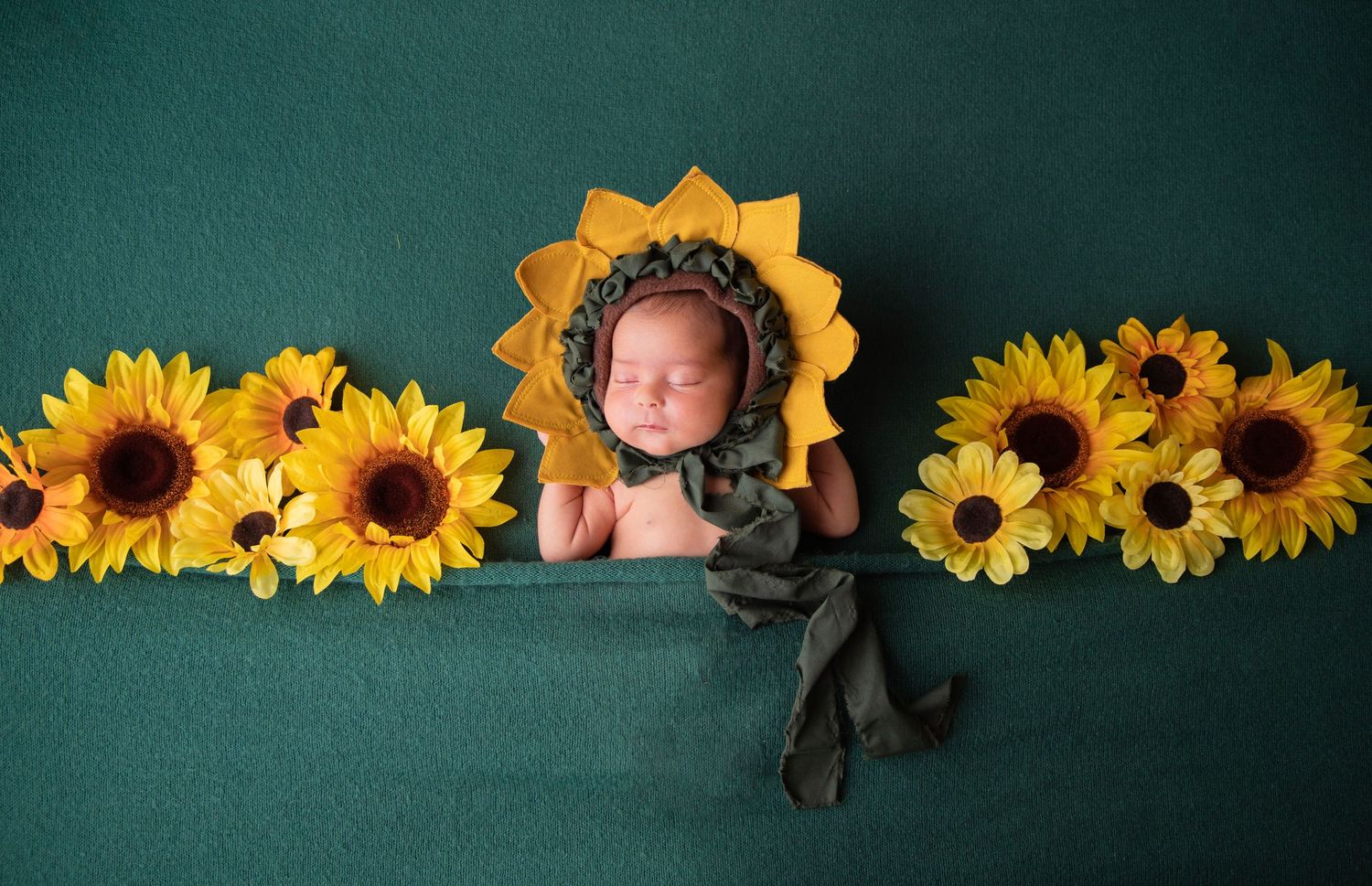 Sleeping newborn baby wearing a sunflower bonnet surrounded by yellow sunflowers on a green backdrop.