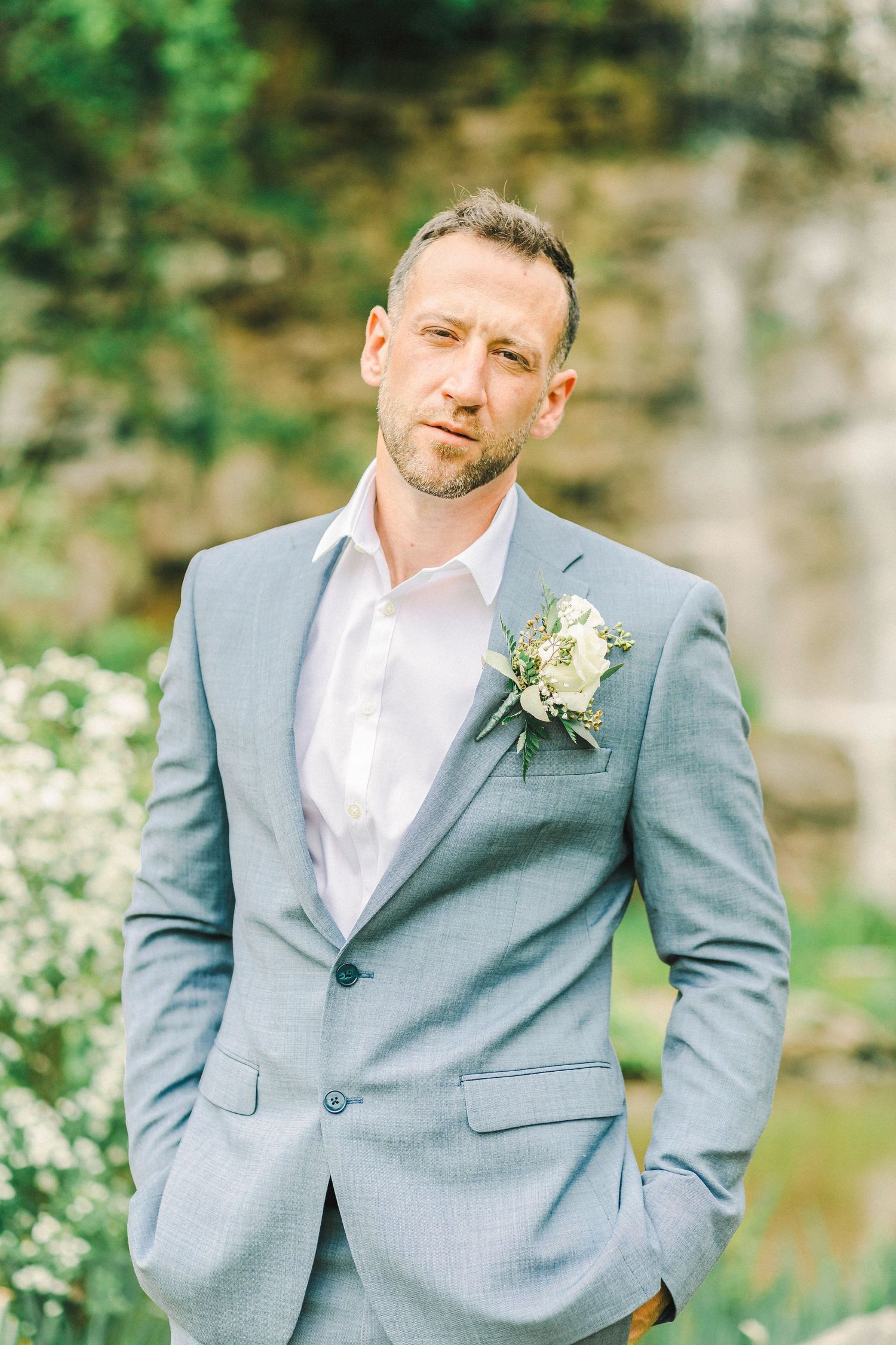 A groom in a light gray suit with a white boutonniere poses outdoors against a natural stone background. Pittsburgh wedding Photographer