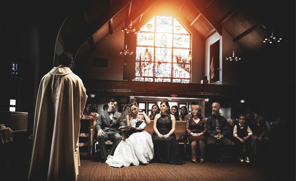 bride and groom sitting in front of a church with priest in the stained glass, in the style of dark black and amber