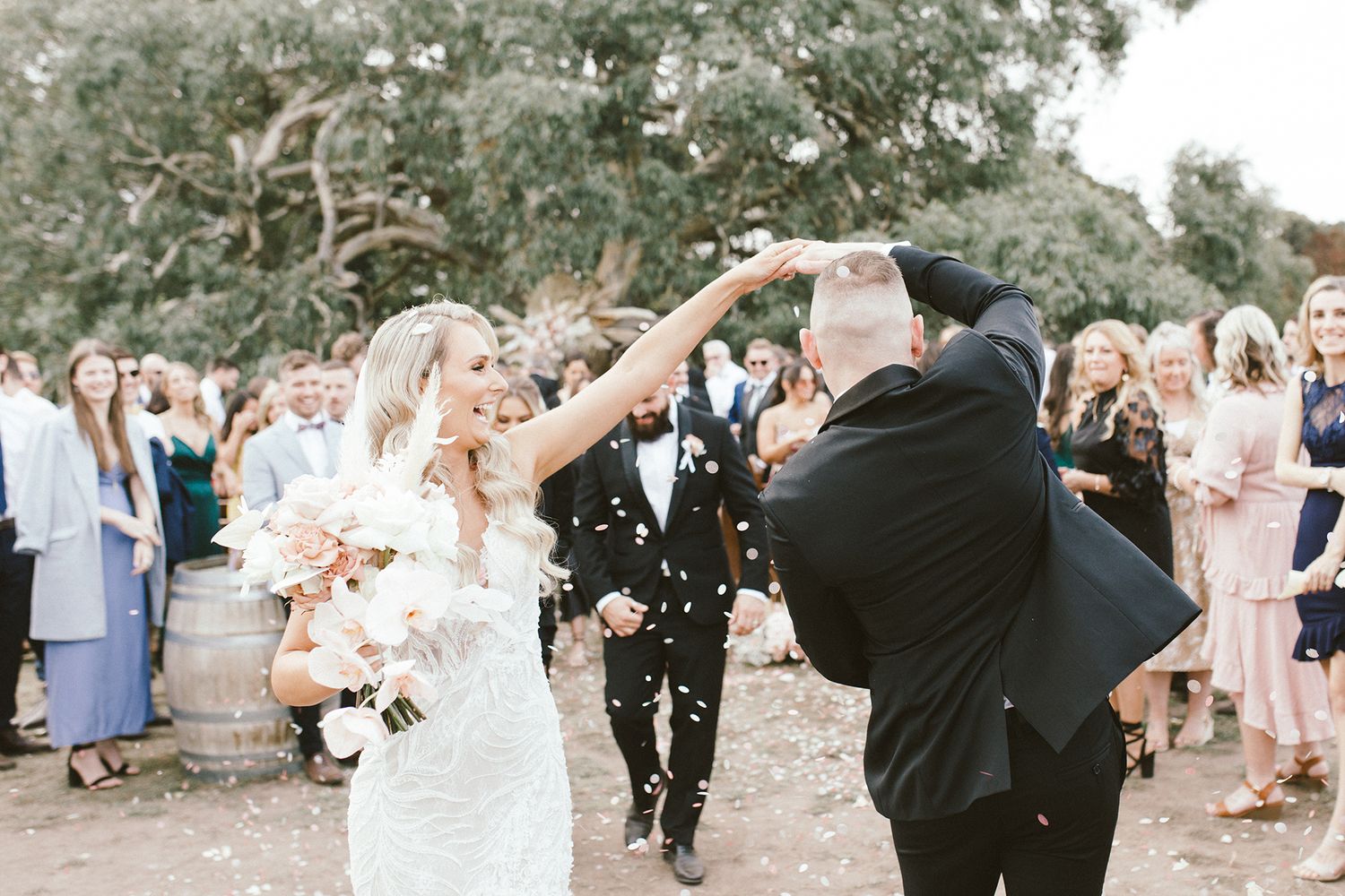 Wedding guests cheer as newlyweds dance and celebrate outdoors beneath trees with confetti flying.