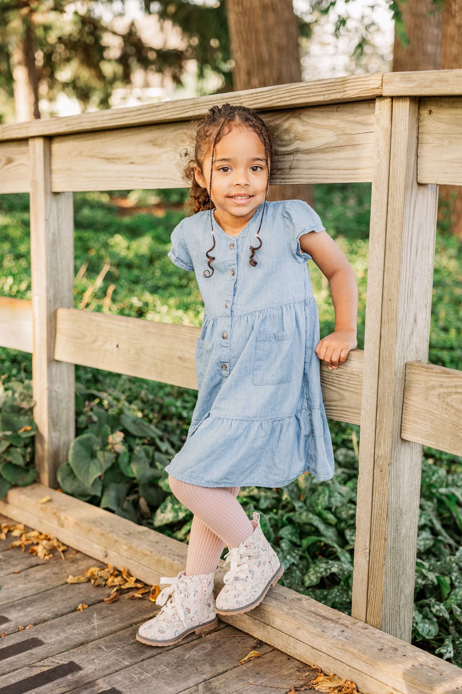Young child in a blue denim dress poses on a wooden bridge with a cheerful expression.