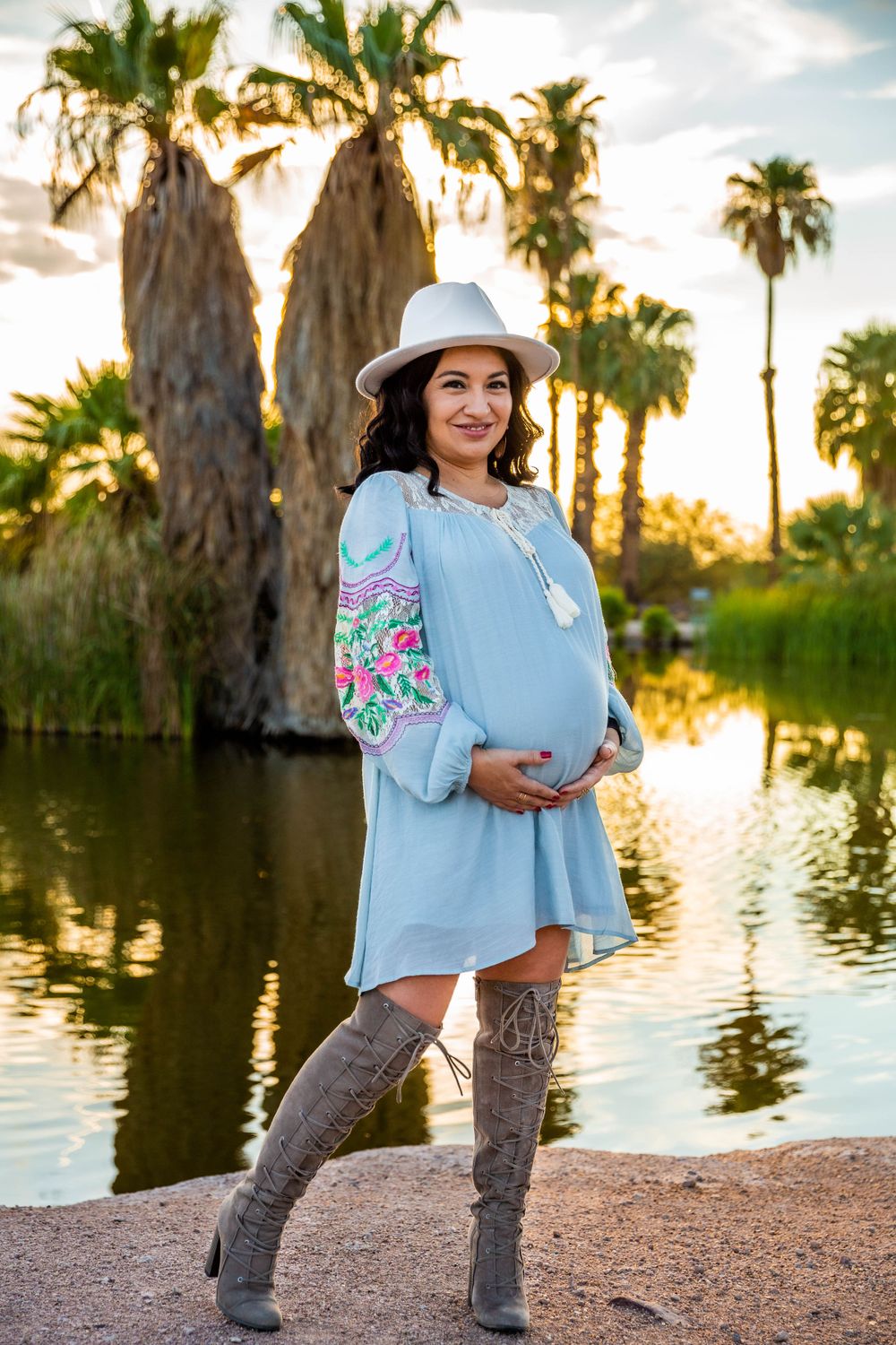Woman in blue dress and hat poses for maternity photo by palm trees and water at sunset.