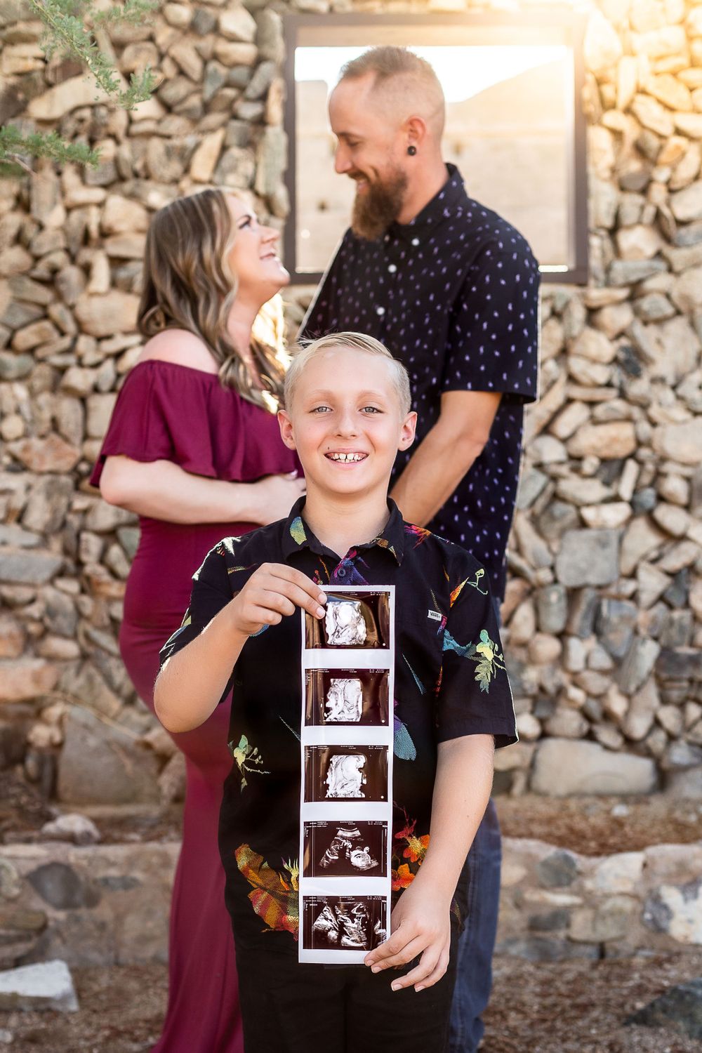 Family pregnancy announcement with child holding ultrasound photo strip in front of stone wall.