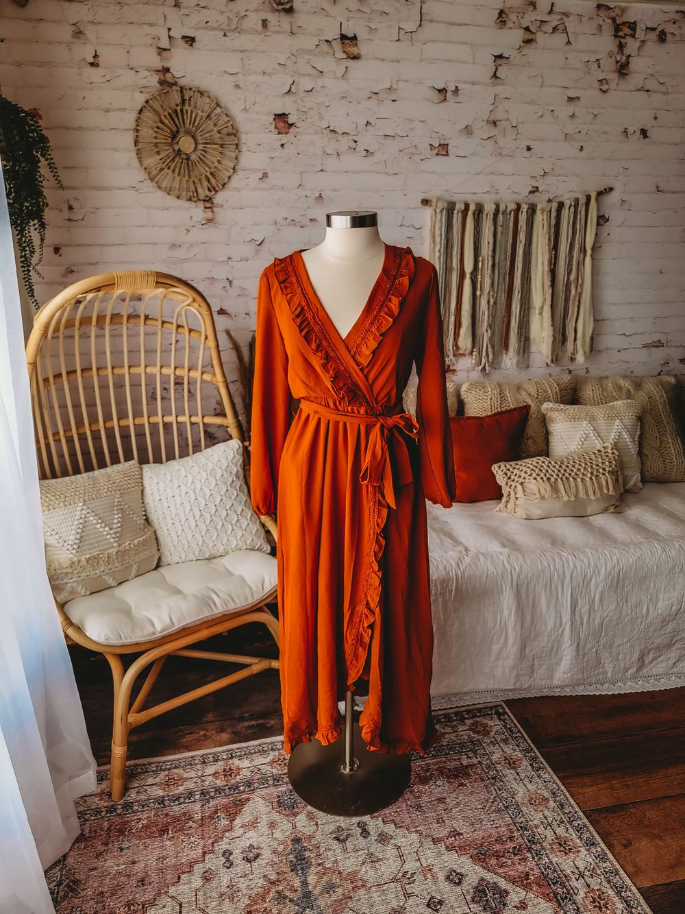 Rust orange long sleeve wrap maxi dress with ruffle details and tie waist on a mannequin in a boho room.