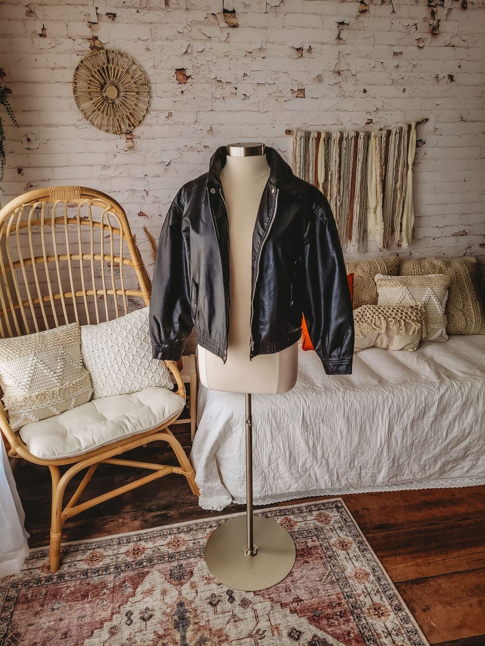 Black faux leather bomber jacket on mannequin in bohemian styled room with rattan chair.