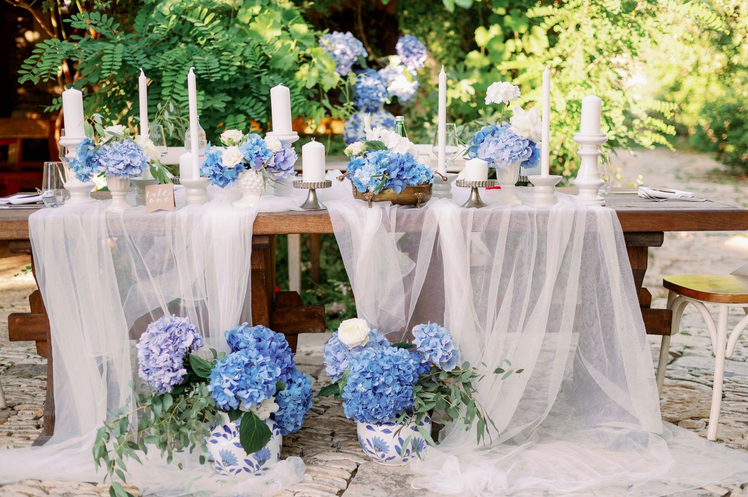 Elegant wedding table setting with blue hydrangeas and white tulle runner on rustic wooden table in garden setting.