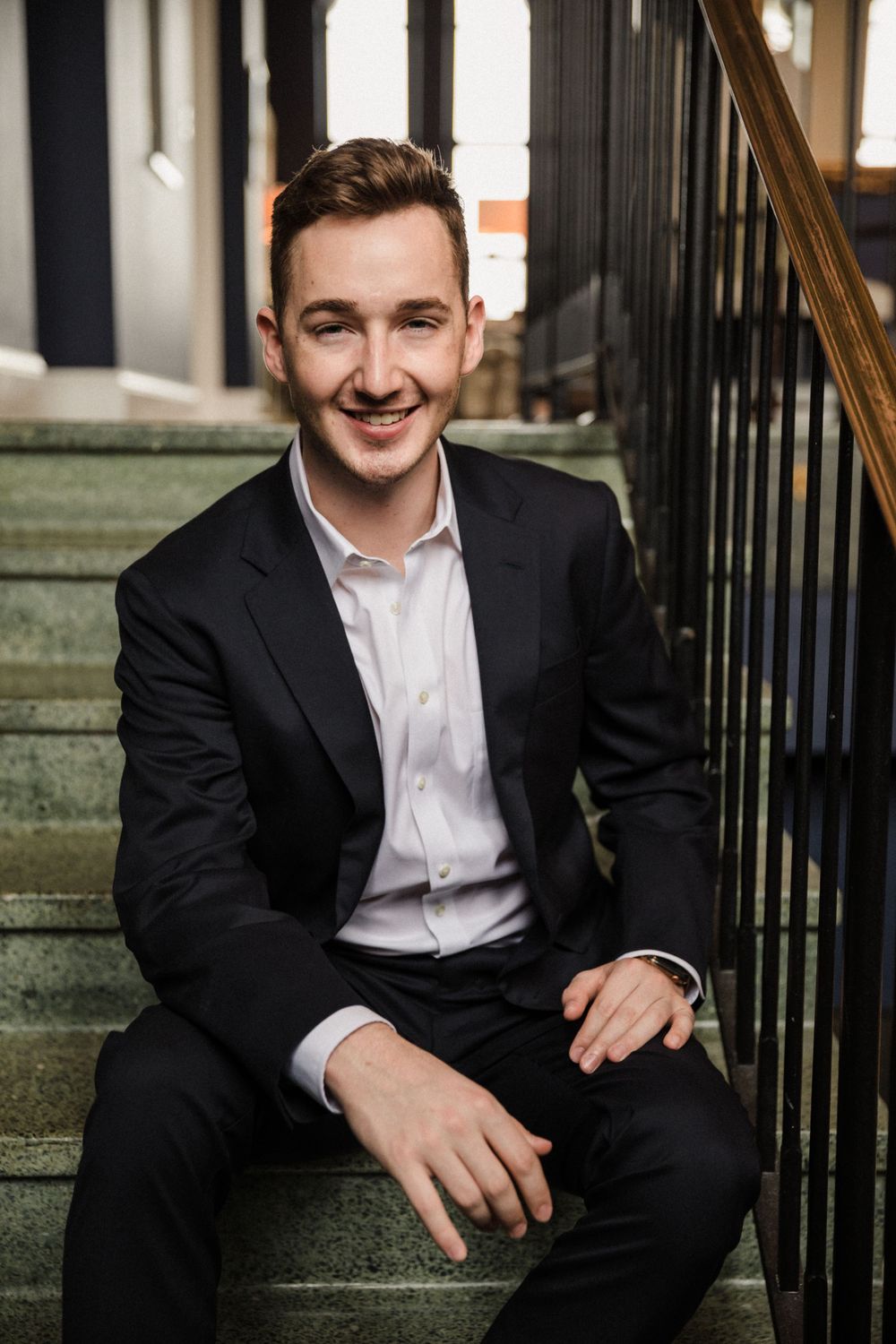 Professional portrait of man in black suit seated on outdoor steps.