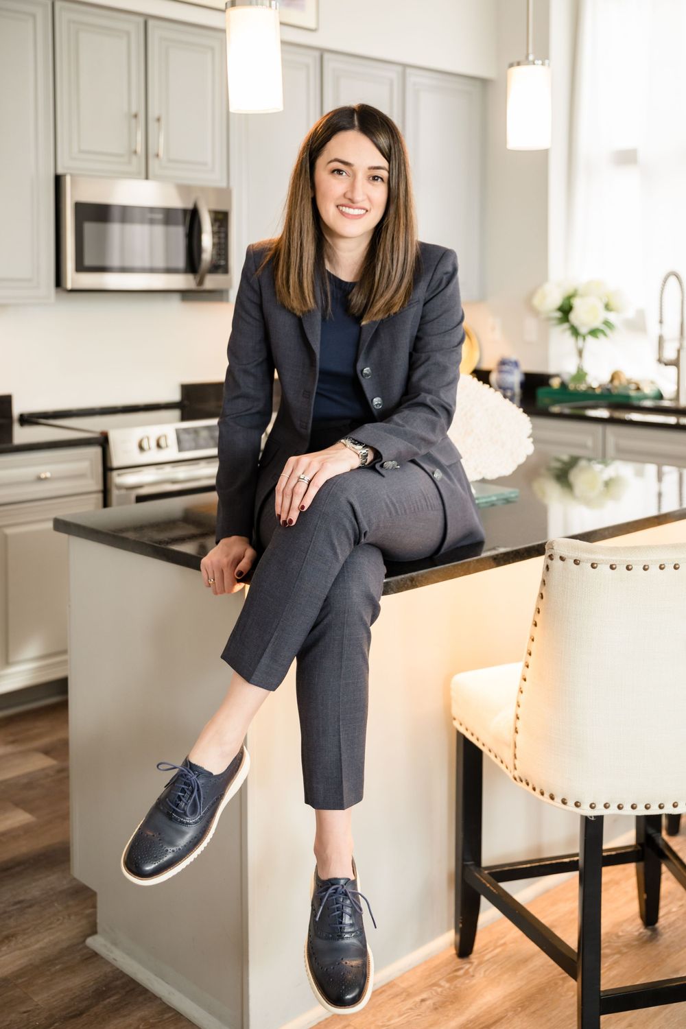 Professional woman in business attire sits on kitchen counter wearing oxford shoes.