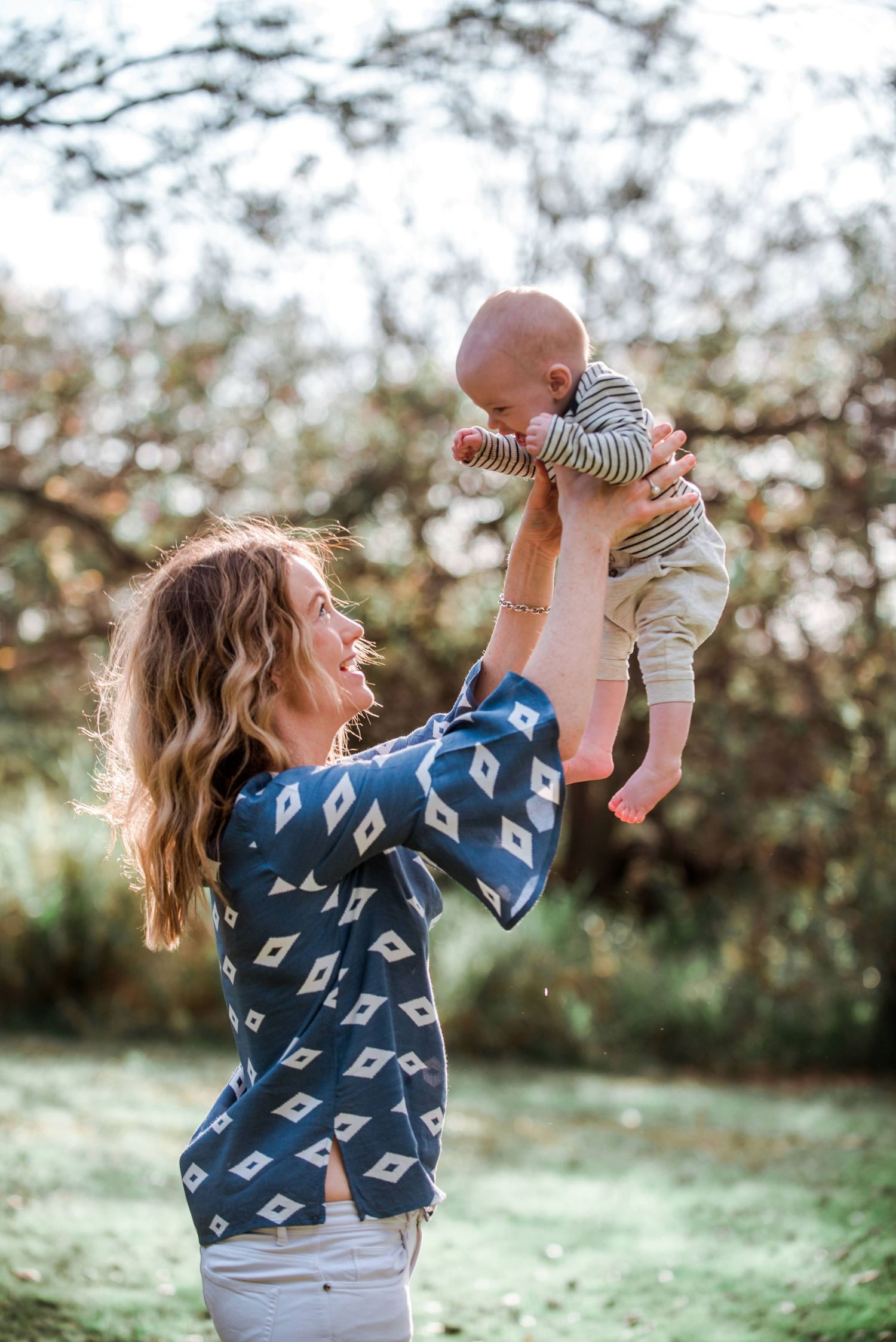 Parent in blue diamond print top lifts giggling baby high in the air outdoors.