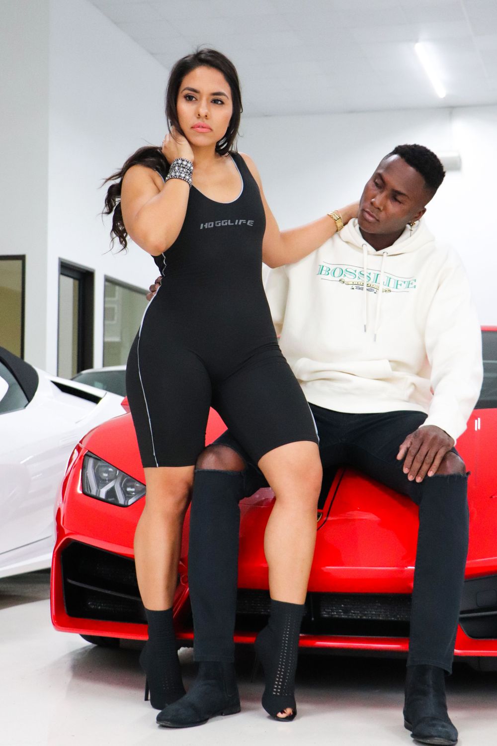 Two models pose together on a red sports car in a white showroom.