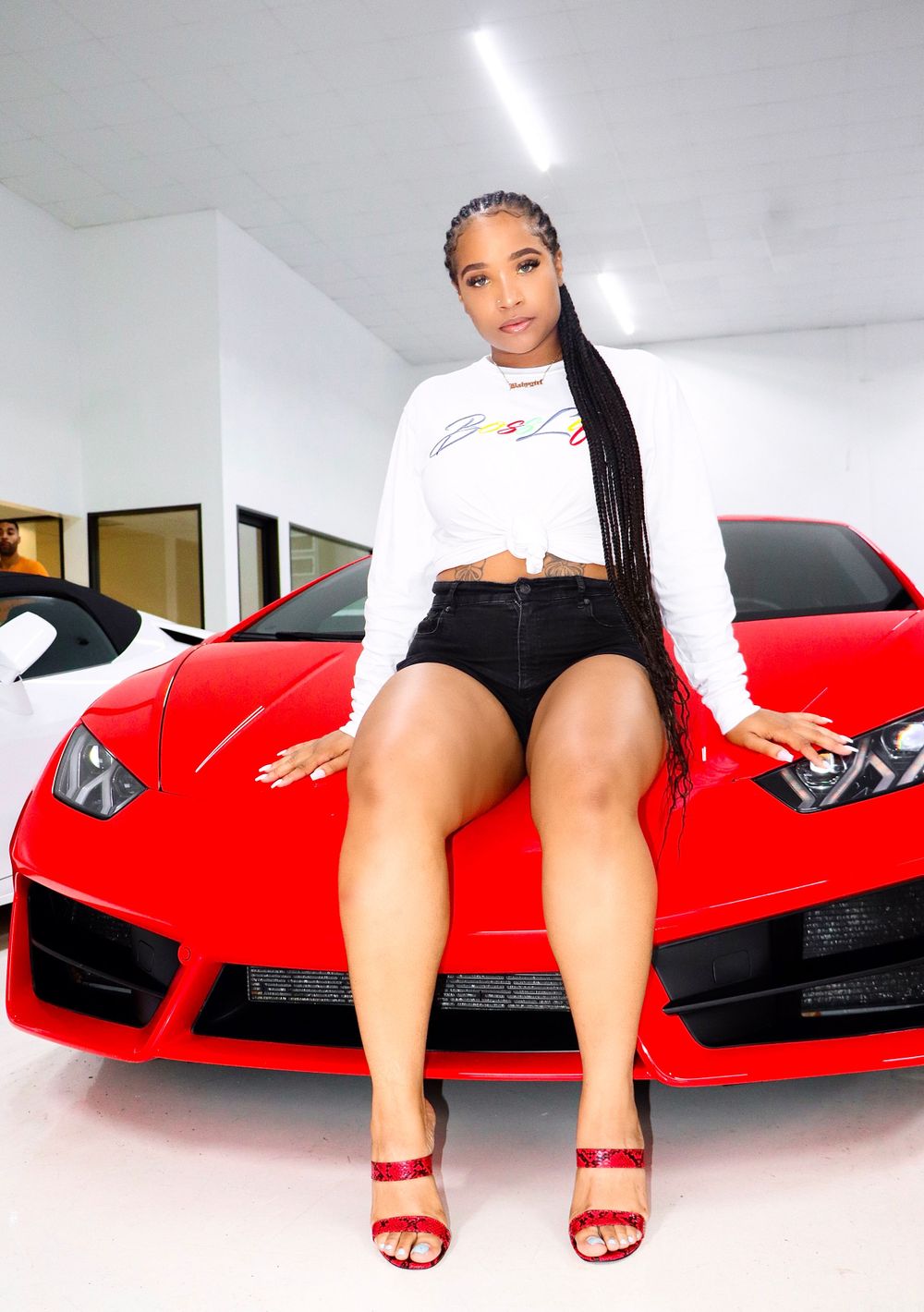 A model poses on the hood of a bright red luxury sports car wearing a white sweater and red sandals.