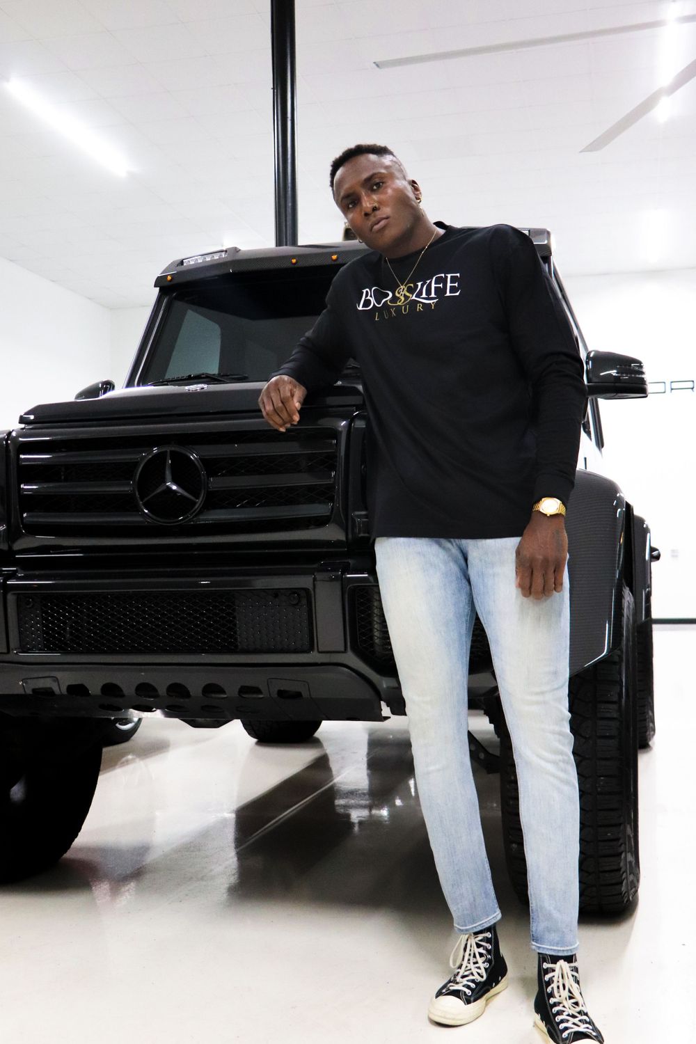 Model in black BOXX4LIFE hoodie leans on black Mercedes G-Wagon in showroom.