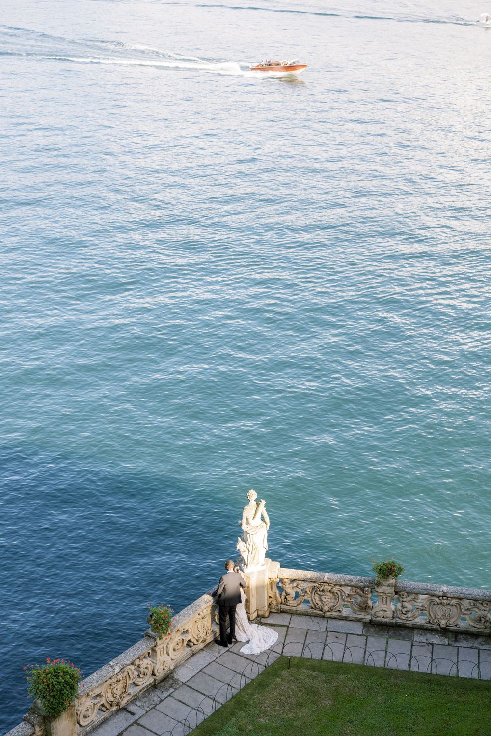 A white statue overlooks the blue Mediterranean Sea from a stone terrace with a boat visible in the distance.