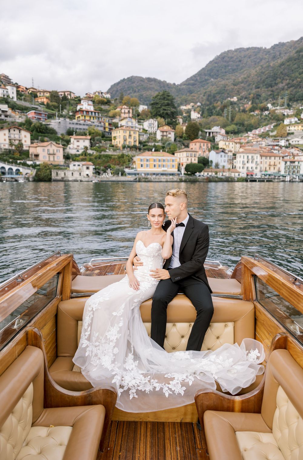 Newlyweds in formal attire on wooden boat against Italian coastal village backdrop.