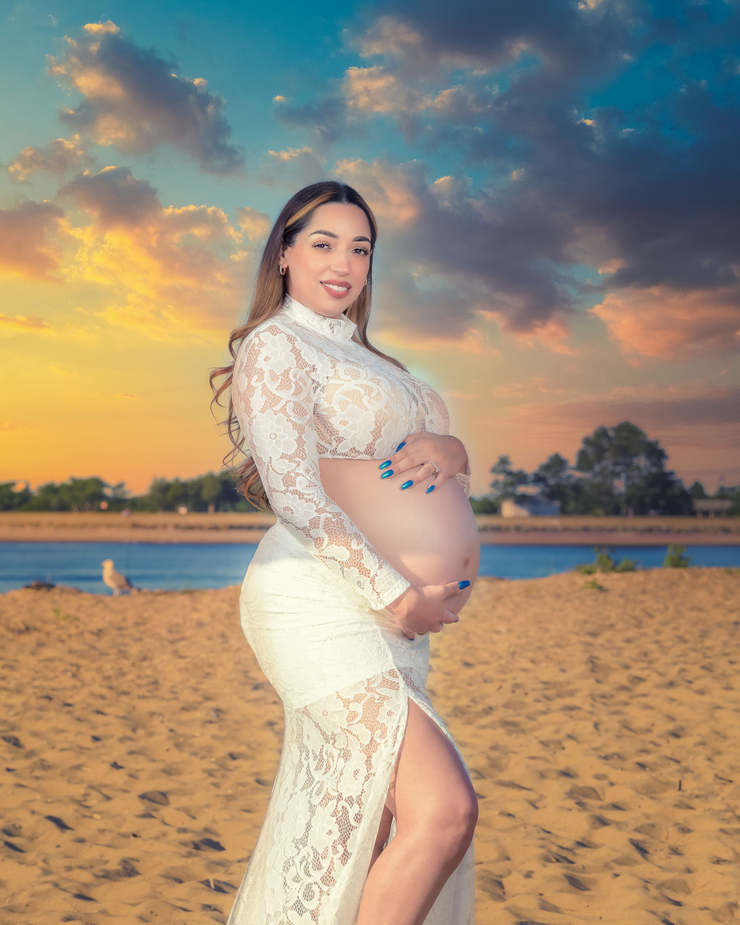 Maternity beach photoshoot at sunset wearing white lace dress on sandy shore.