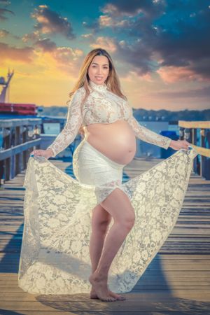 Beachside maternity session with lace dress and dramatic sunset lighting.