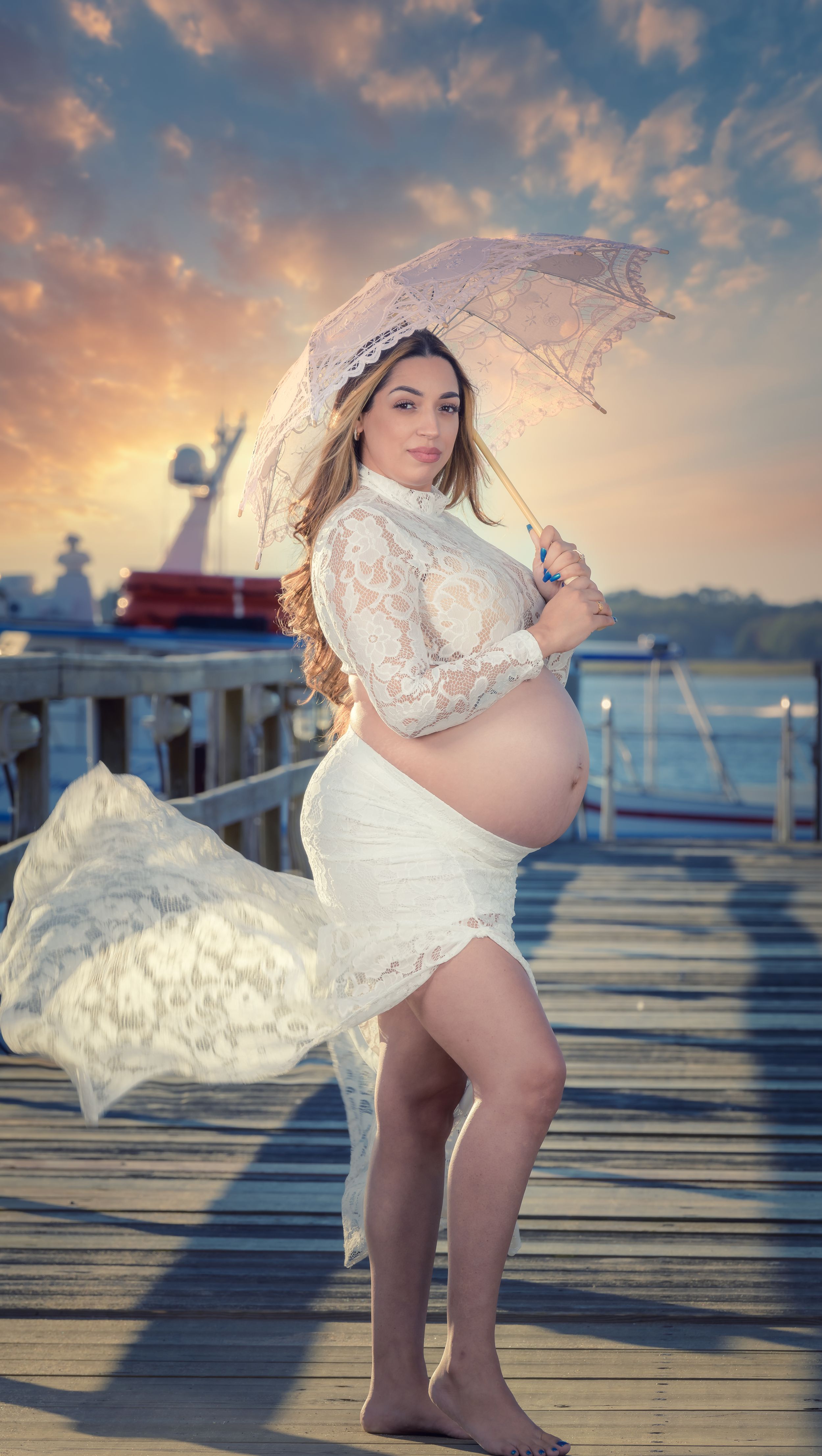 Maternity photos on wooden pier at sunset with flowing white lace dress.