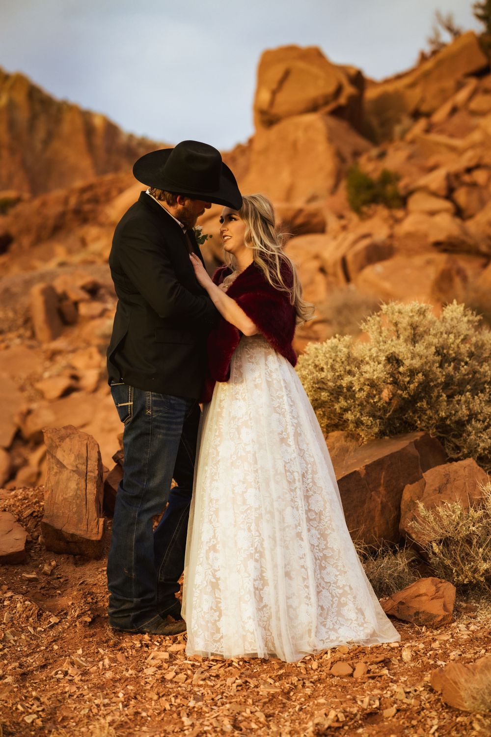 Bride in white lace gown and groom in black cowboy hat share a romantic moment in red rock desert.