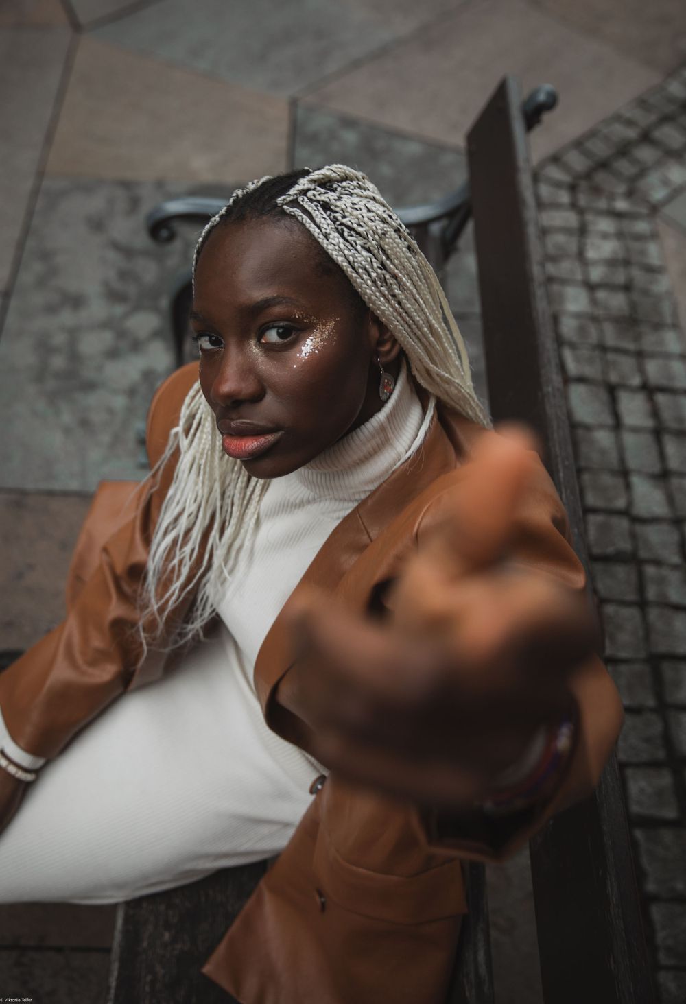 Fashion portrait featuring white top and tan coat with blonde braided hair against urban backdrop.